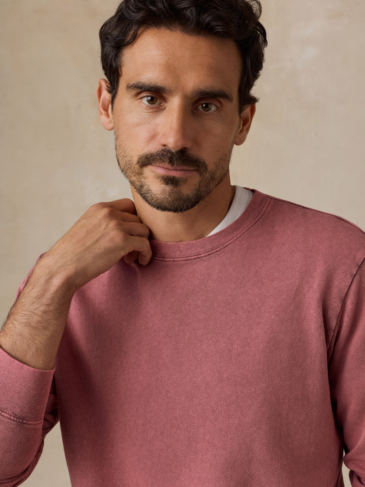 A close-up of a man wearing a dusty rose crewneck sweatshirt over a white undershirt, set against a neutral beige background., image 4 of 7