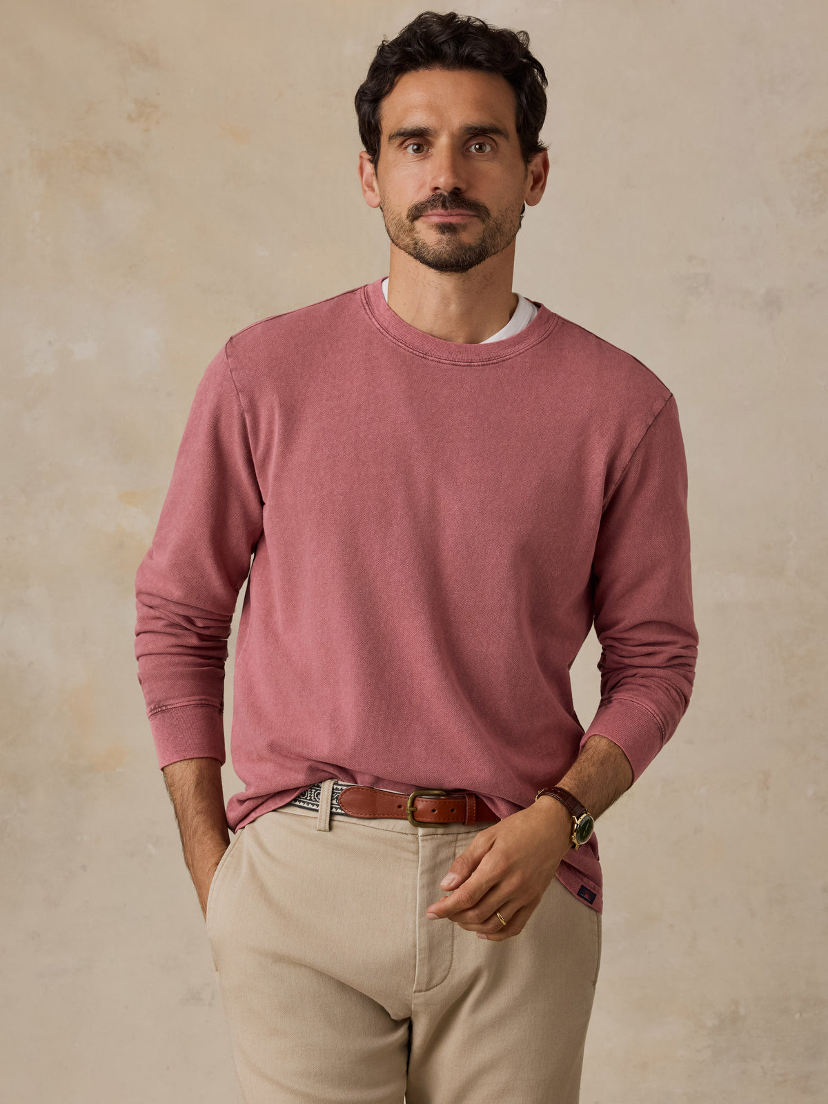 A man wearing a dusty rose long-sleeve crewneck sweater, beige trousers, and a brown leather belt, standing against a textured beige background., image 3 of 7
