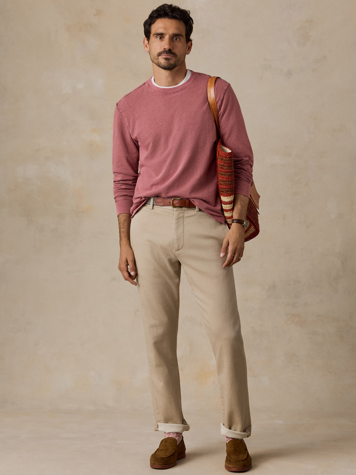 A man stands against a textured beige studio backdrop wearing a dusty rose long-sleeve crewneck sweater, beige trousers, brown suede loafers, and carrying a brown leather shoulder bag., image 6 of 7