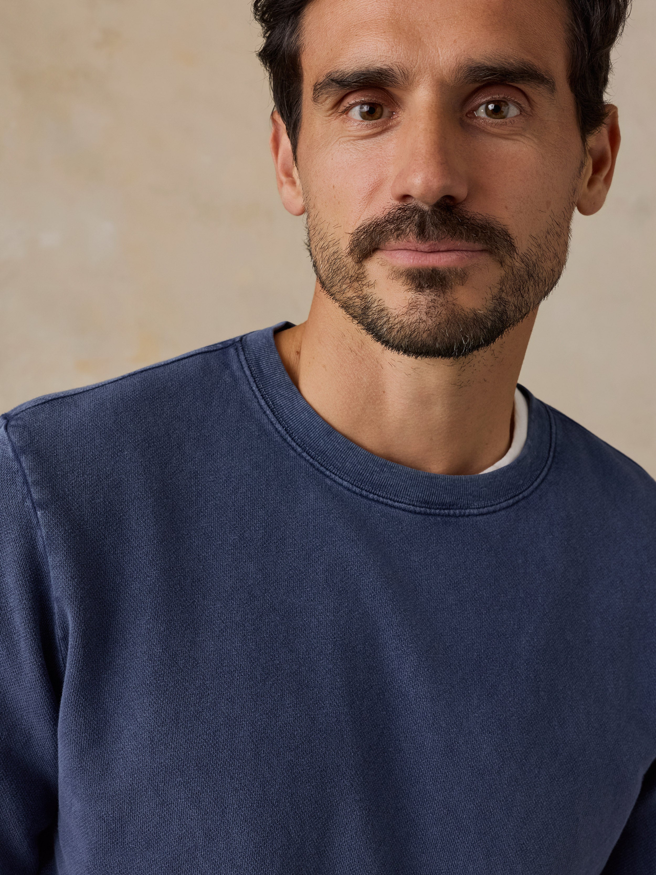 A close-up shot of a man wearing a navy blue crewneck sweatshirt, showing the fabric texture and collar detail against a neutral beige background.