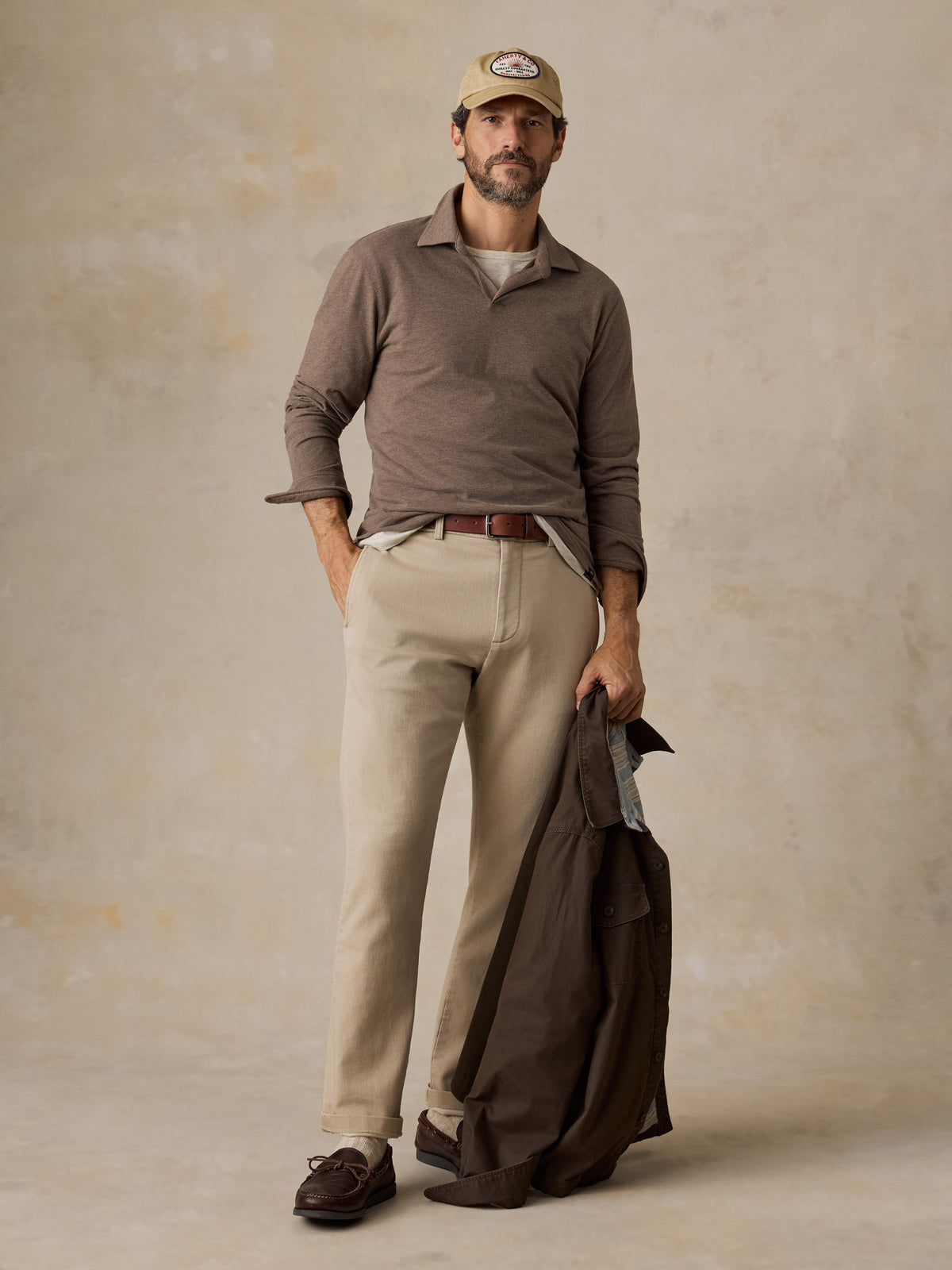 A man wearing a brown polo shirt, beige chino pants, beige hat, and brown jacket, image 6 of 6