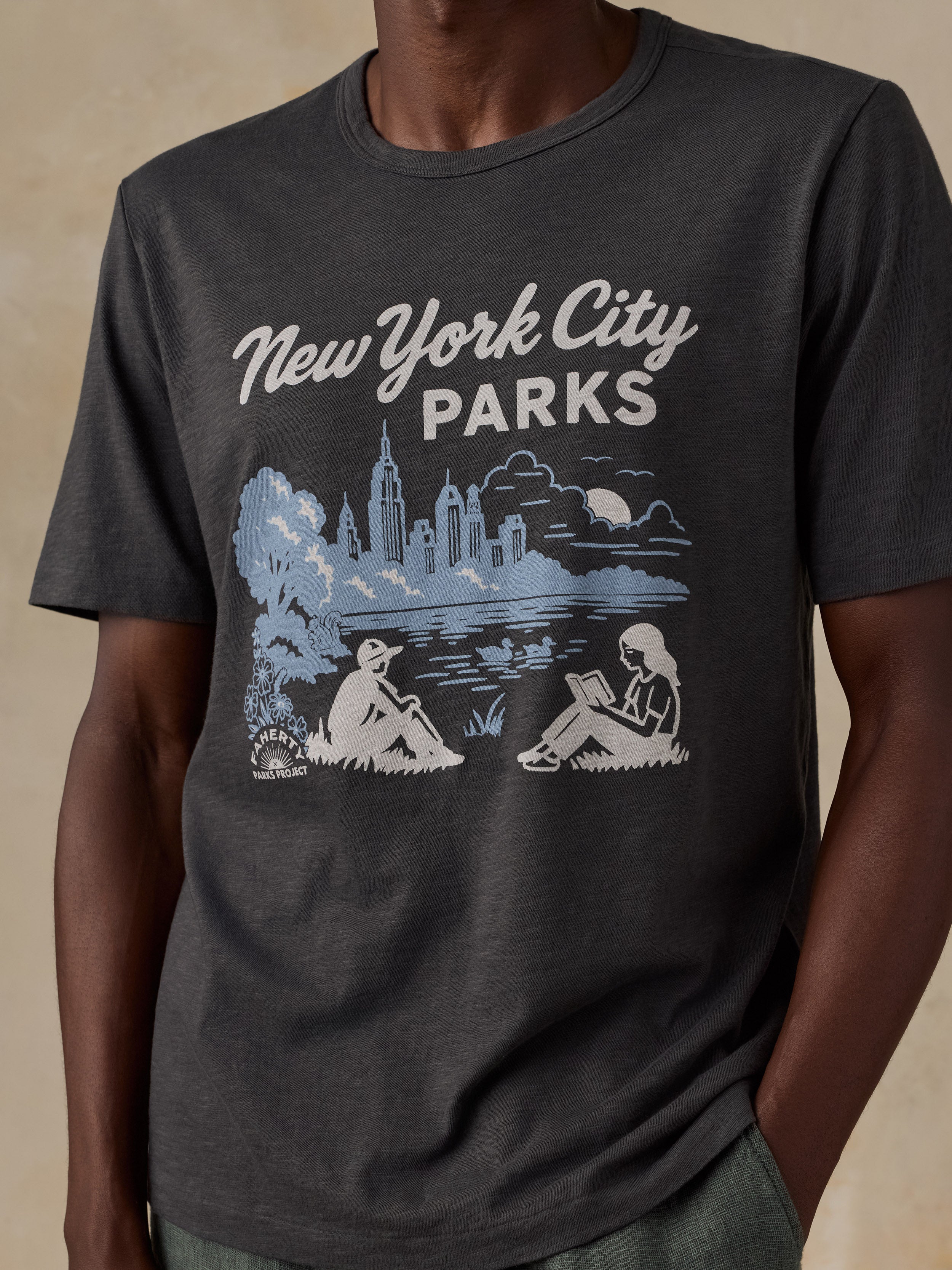 A man wearing a dark charcoal gray t-shirt with a light blue and white graphic print that reads 'New York City Parks' above an illustration of the skyline and people sitting in a park.