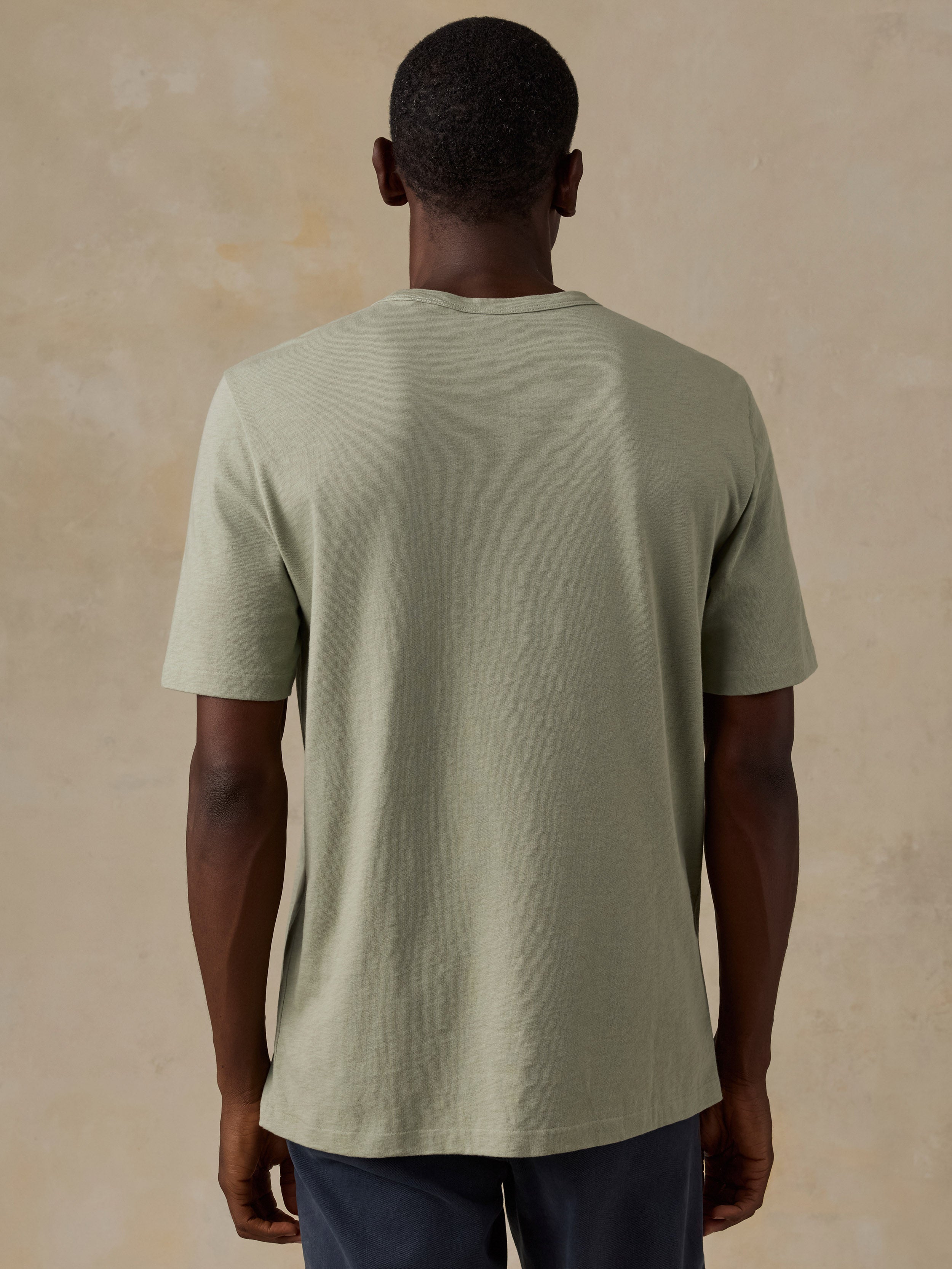 A man viewed from behind wearing a light sage green short-sleeve t-shirt and dark blue pants, standing against a textured beige background.