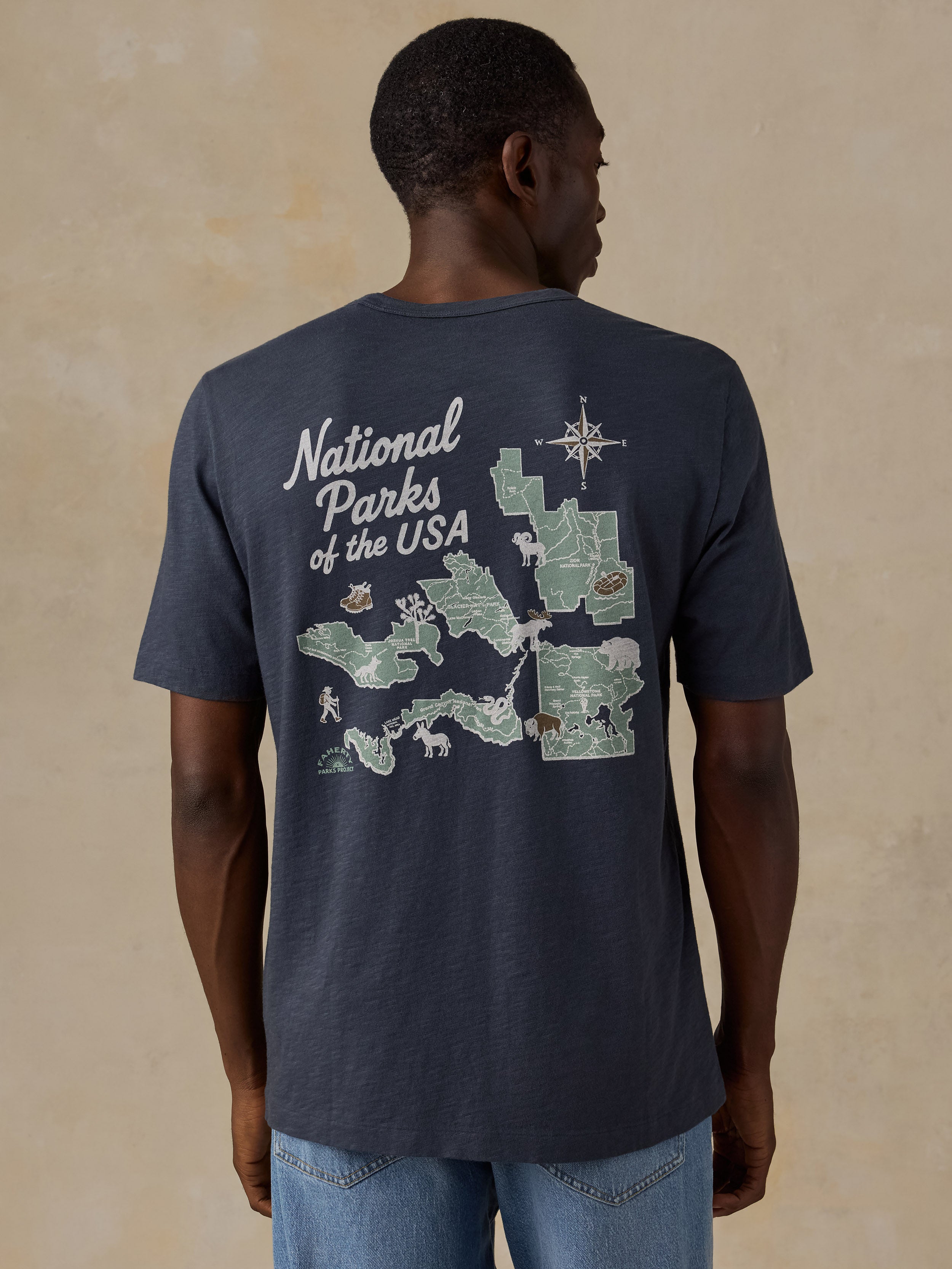 A man wearing a navy blue t-shirt with a large back graphic of a map of National Parks of the USA, shown from the back against a textured beige background.