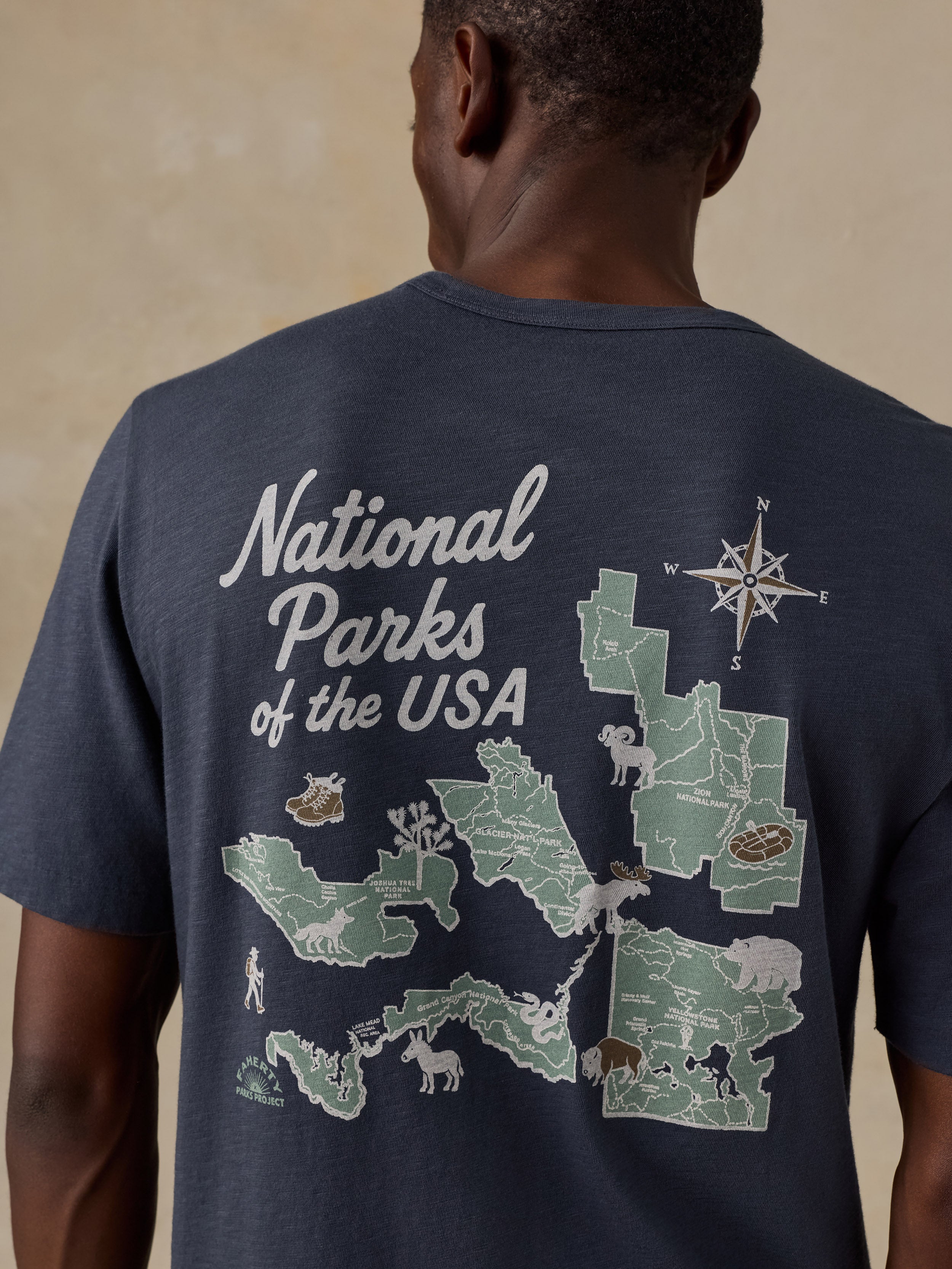 A man viewed from behind wearing a navy blue t-shirt with a large graphic print on the back. The graphic features a map of National Parks of the USA in light green and white, complete with a compass rose and various animal illustrations.