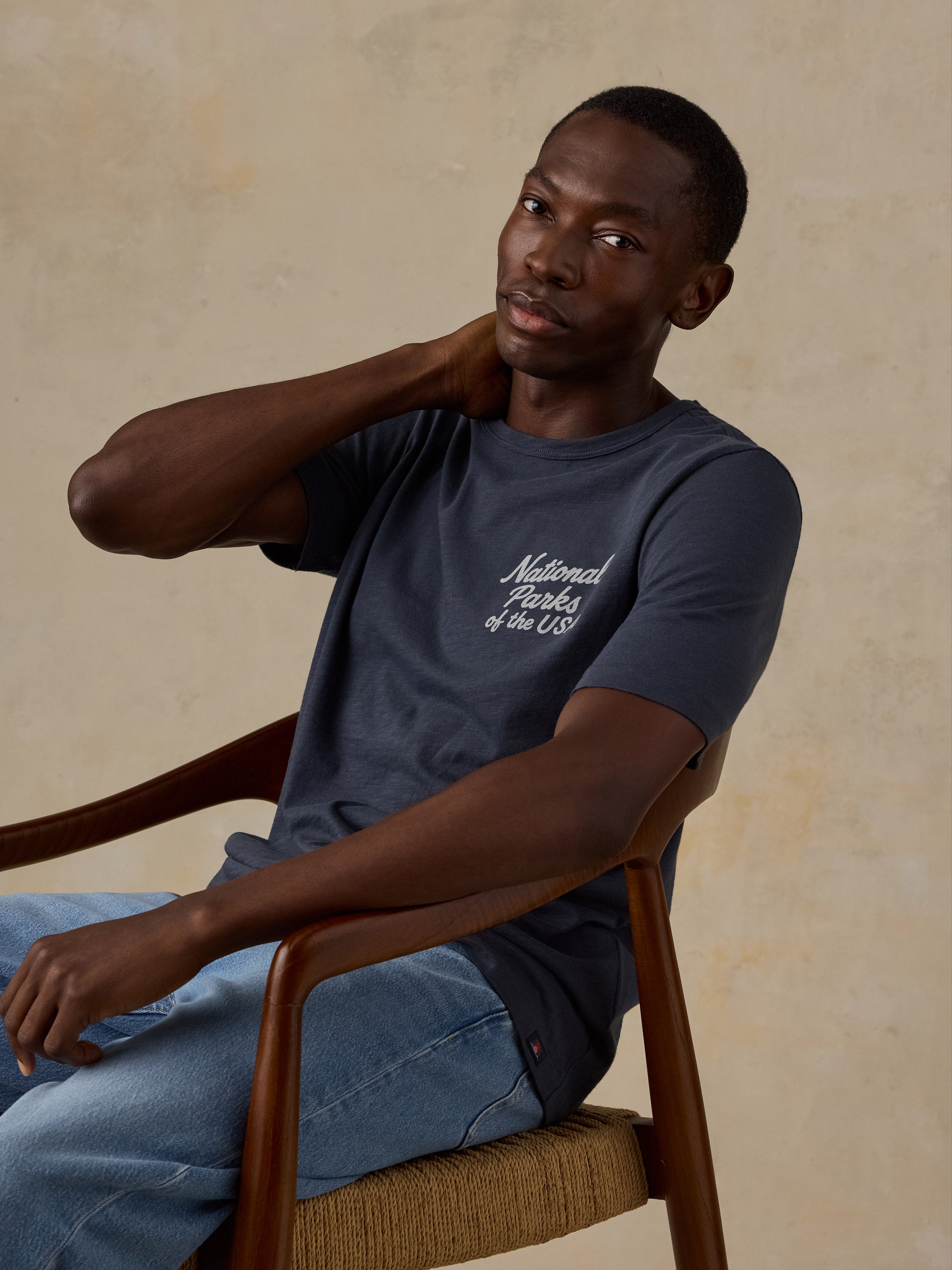 A man wearing a navy blue t-shirt with white cursive text reading 'National Parks of the US' sits in a wooden armchair against a textured beige background.