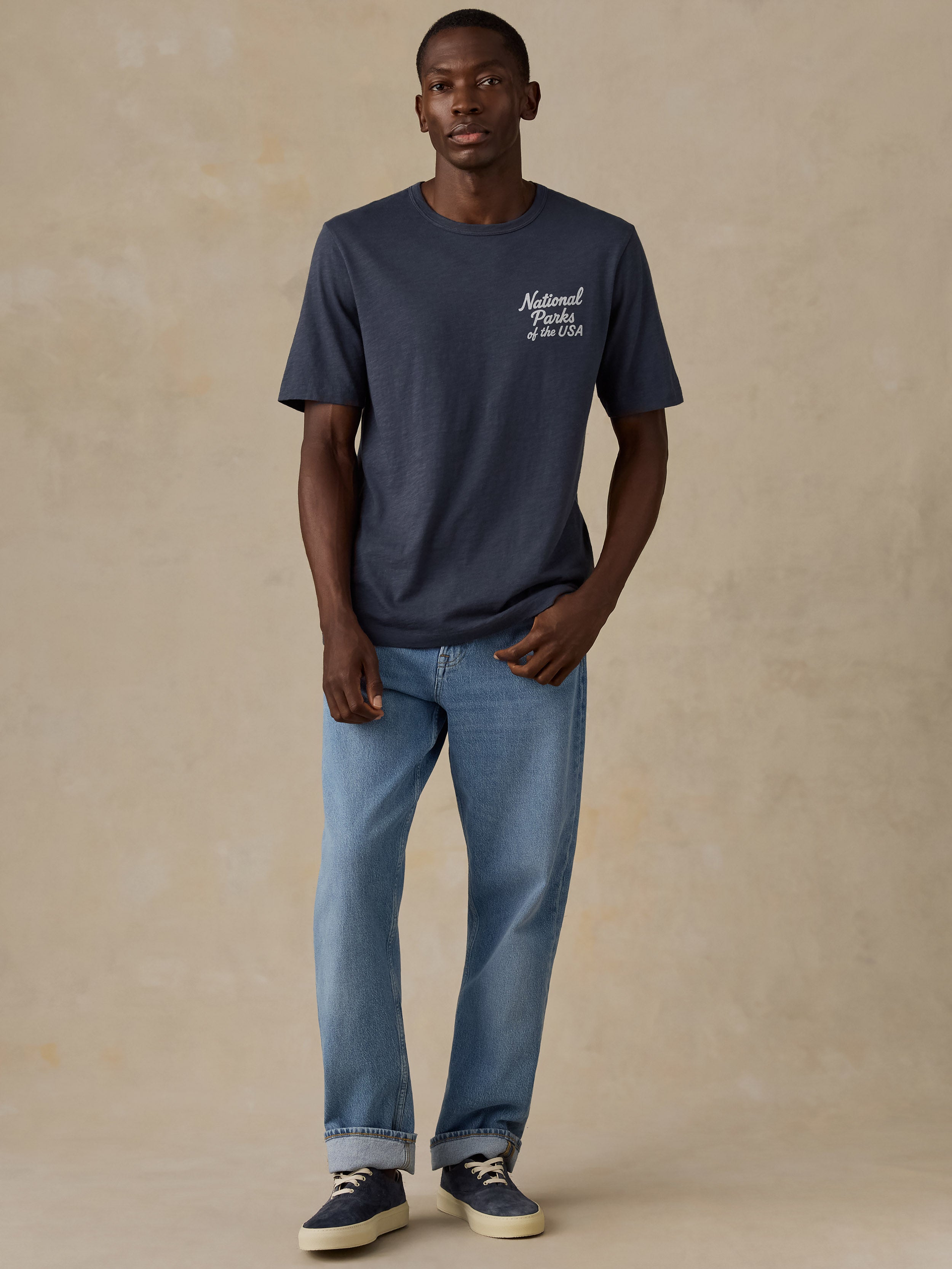 A man modeling a navy blue crewneck t-shirt with a white 'National Parks of the USA' graphic on the left chest, paired with light blue jeans and navy sneakers, against a textured beige studio backdrop.