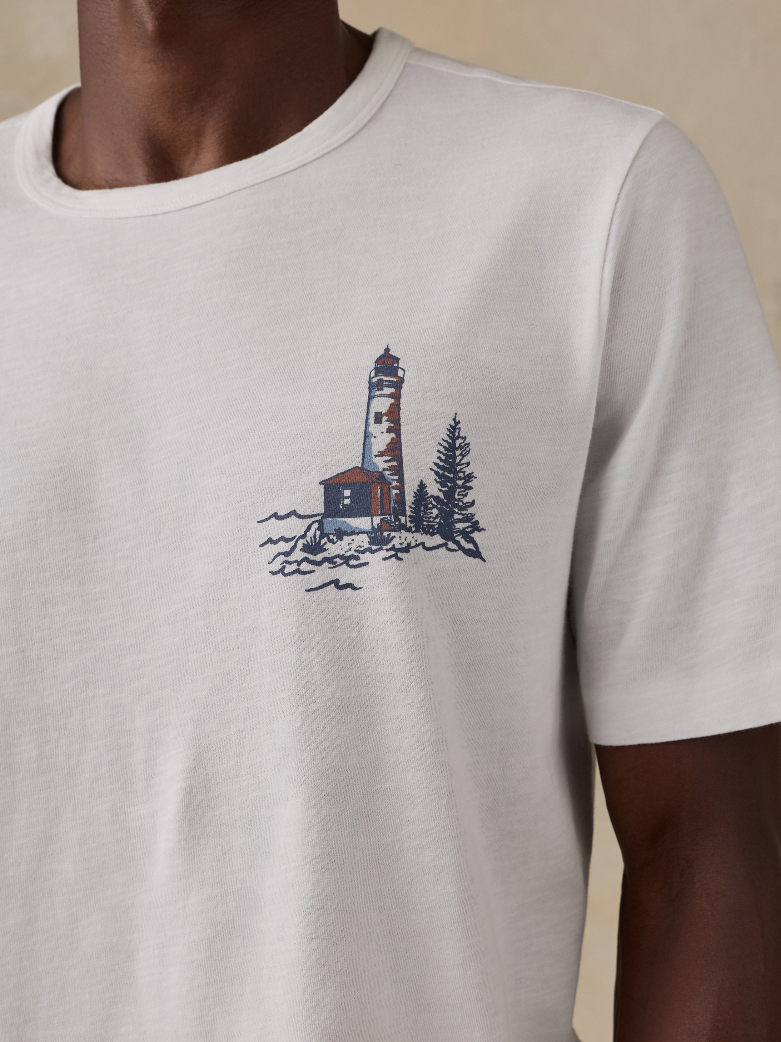 A close-up of a man wearing a white crew-neck t-shirt with a printed graphic of a lighthouse, house, and pine tree on the left chest.