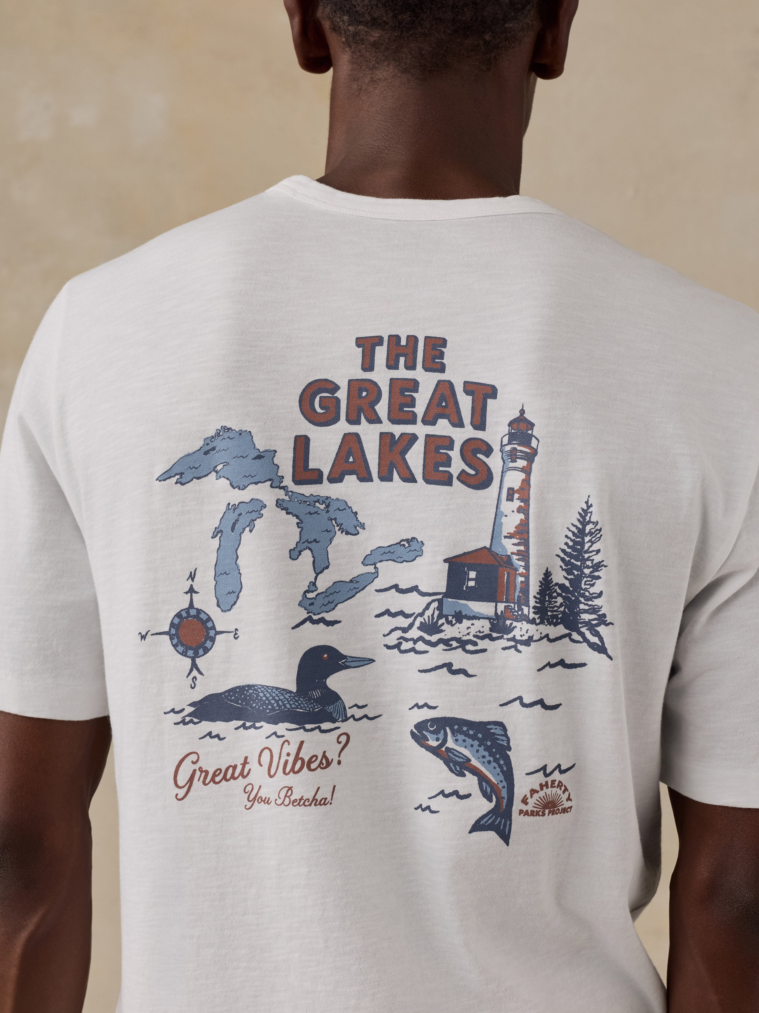 A man viewed from behind wearing a cream-colored t-shirt with a large graphic print on the back featuring the text 'THE GREAT LAKES' and illustrations of Michigan, a lighthouse, a loon, a fish, and the phrase 'Great Vibes You Brought!'.