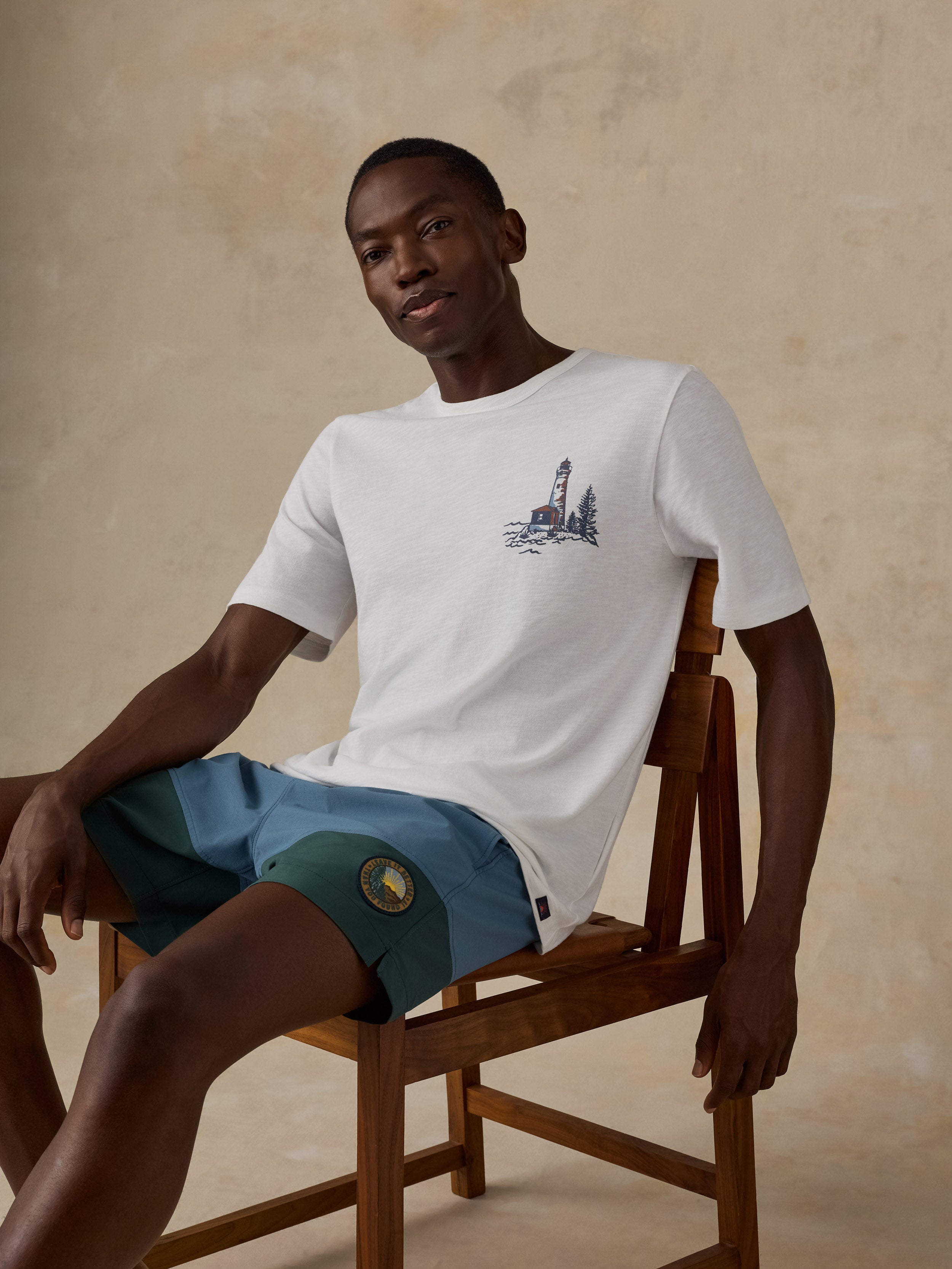 A man sitting on a wooden chair wearing a white crew-neck t-shirt with a small navy blue lighthouse graphic on the left chest, paired with teal shorts, against a textured beige studio background.