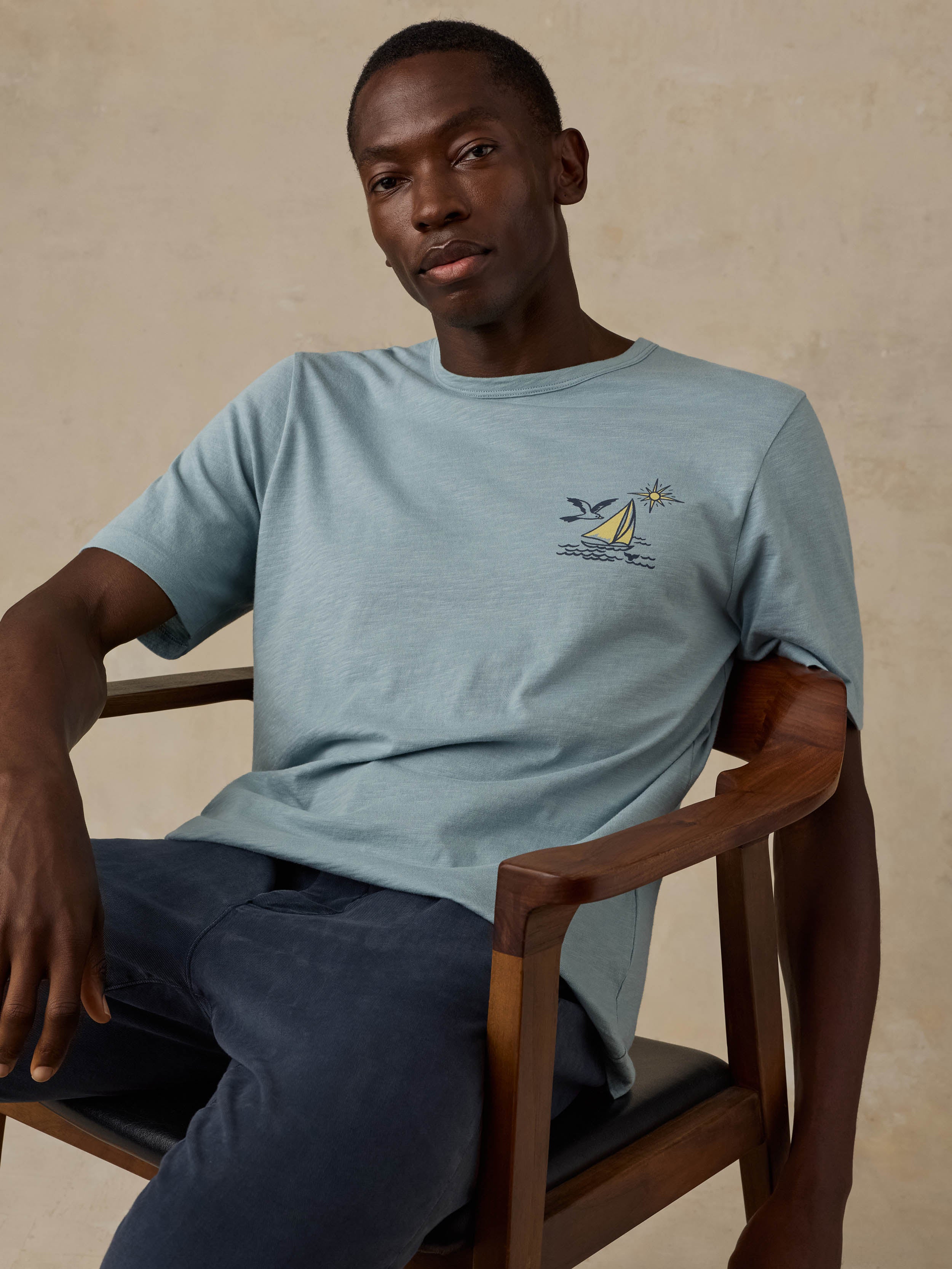 A man sitting in a wooden chair wearing a light blue crew-neck t-shirt with a small sailboat graphic on the left chest, paired with dark blue pants, against a beige background