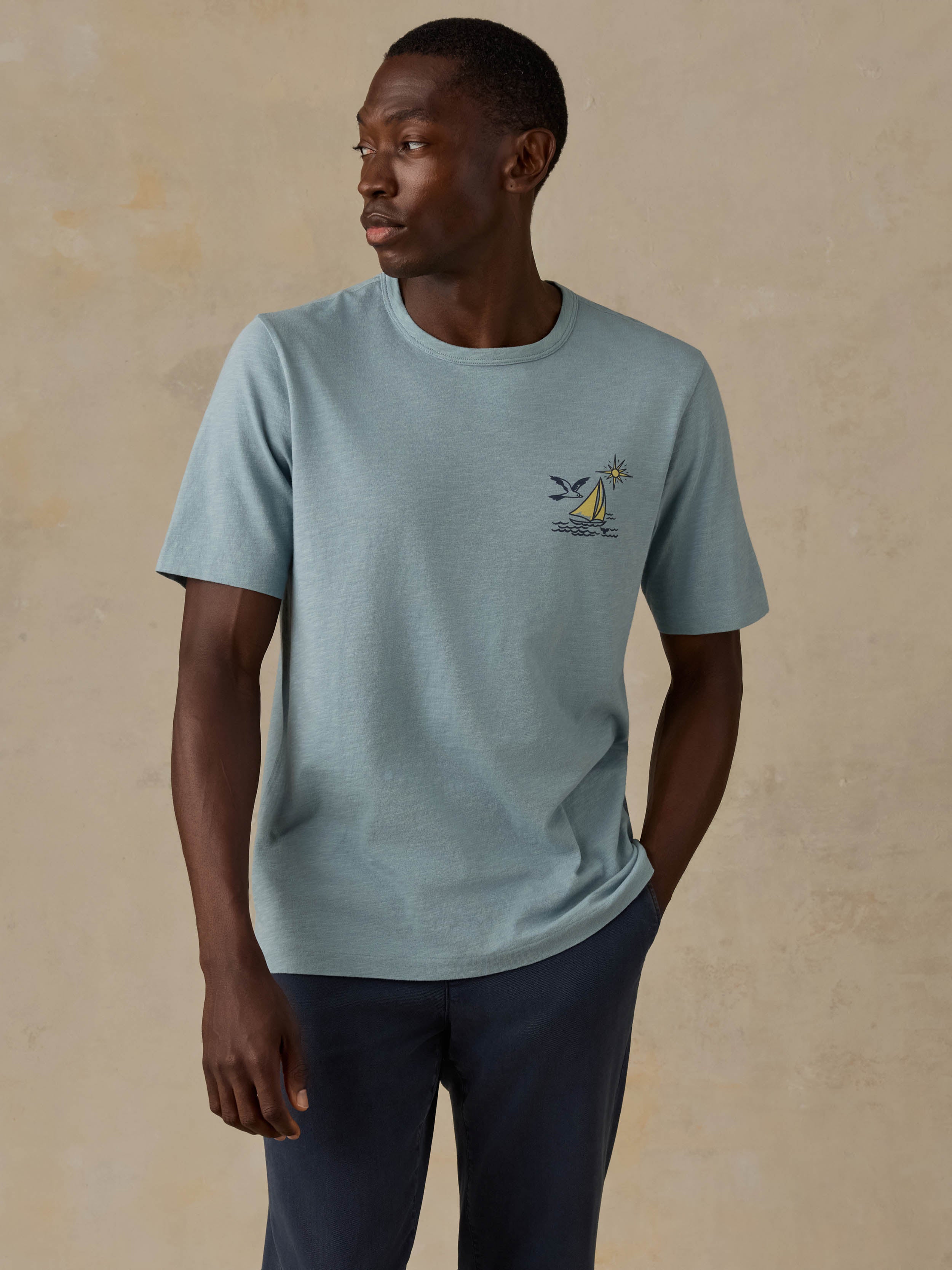 Man wearing a blue t-shirt with a sailboat graphic on the left chest and blue pants against a beige background