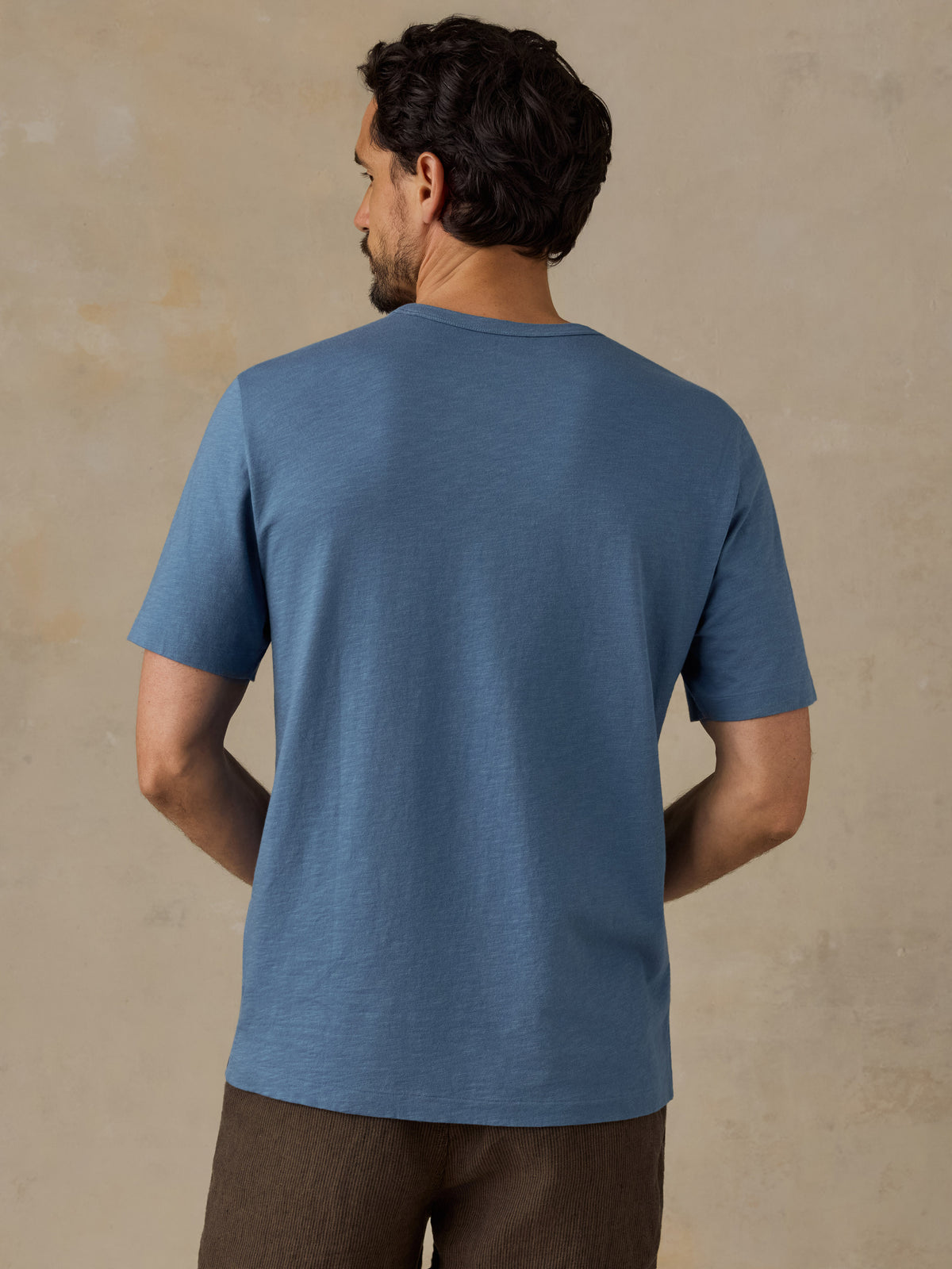 A man viewed from the back wearing a heathered slate blue short-sleeve t-shirt and brown pants, standing against a textured beige studio backdrop., image 3 of 6