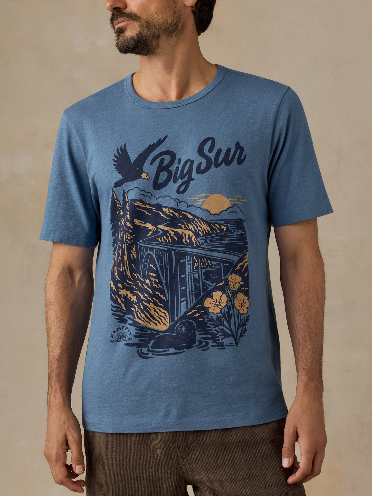 A man wearing a heathered blue crewneck t-shirt with a large graphic print on the front featuring the text 'Big Sur' and an illustration of a cabin, mountains, a bird, and flowers., image 5 of 6