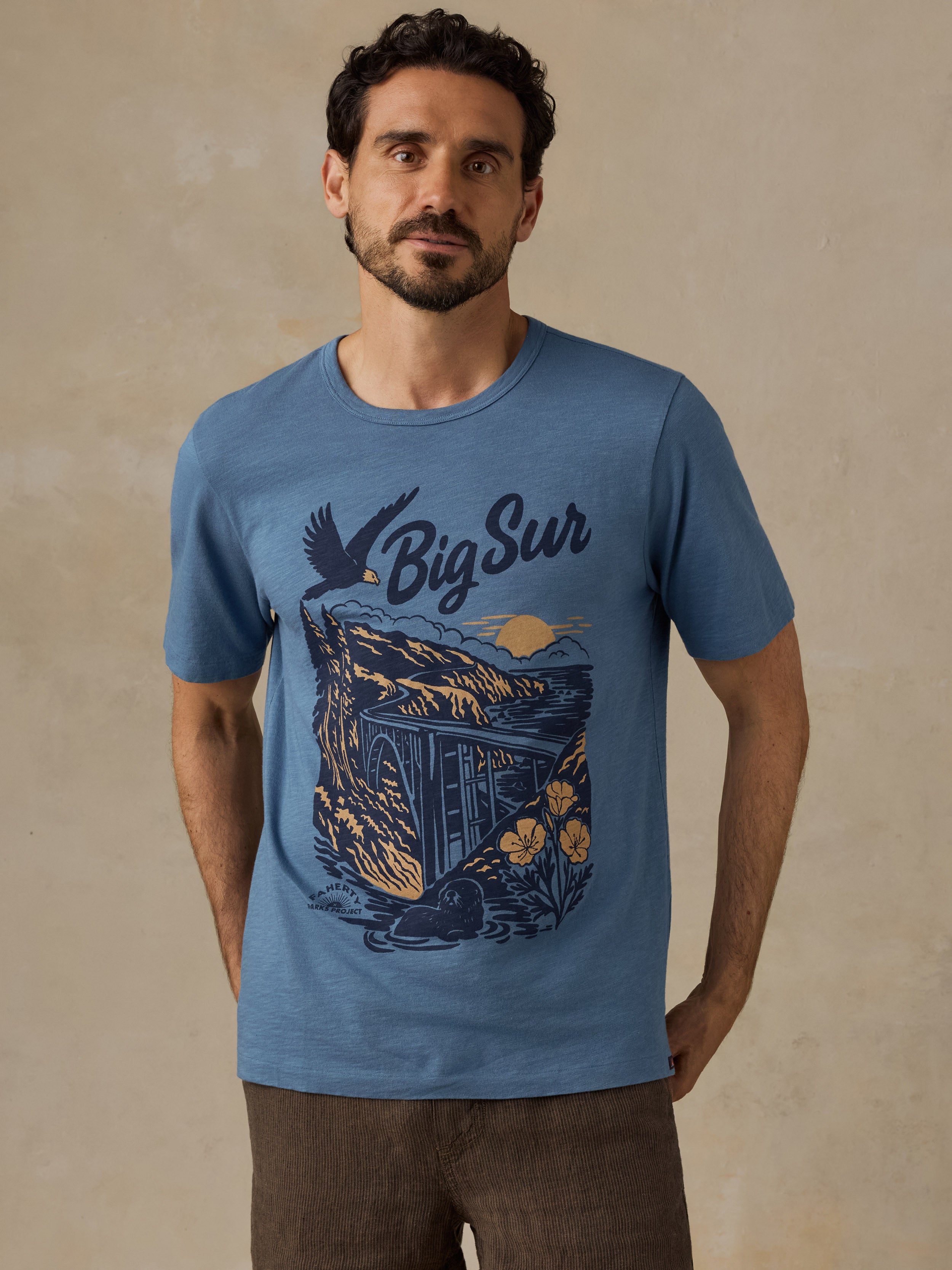 A man wearing a heather blue graphic t-shirt with a 'Big Sur' coastal illustration in navy and yellow, paired with brown pants, standing against a textured beige wall.