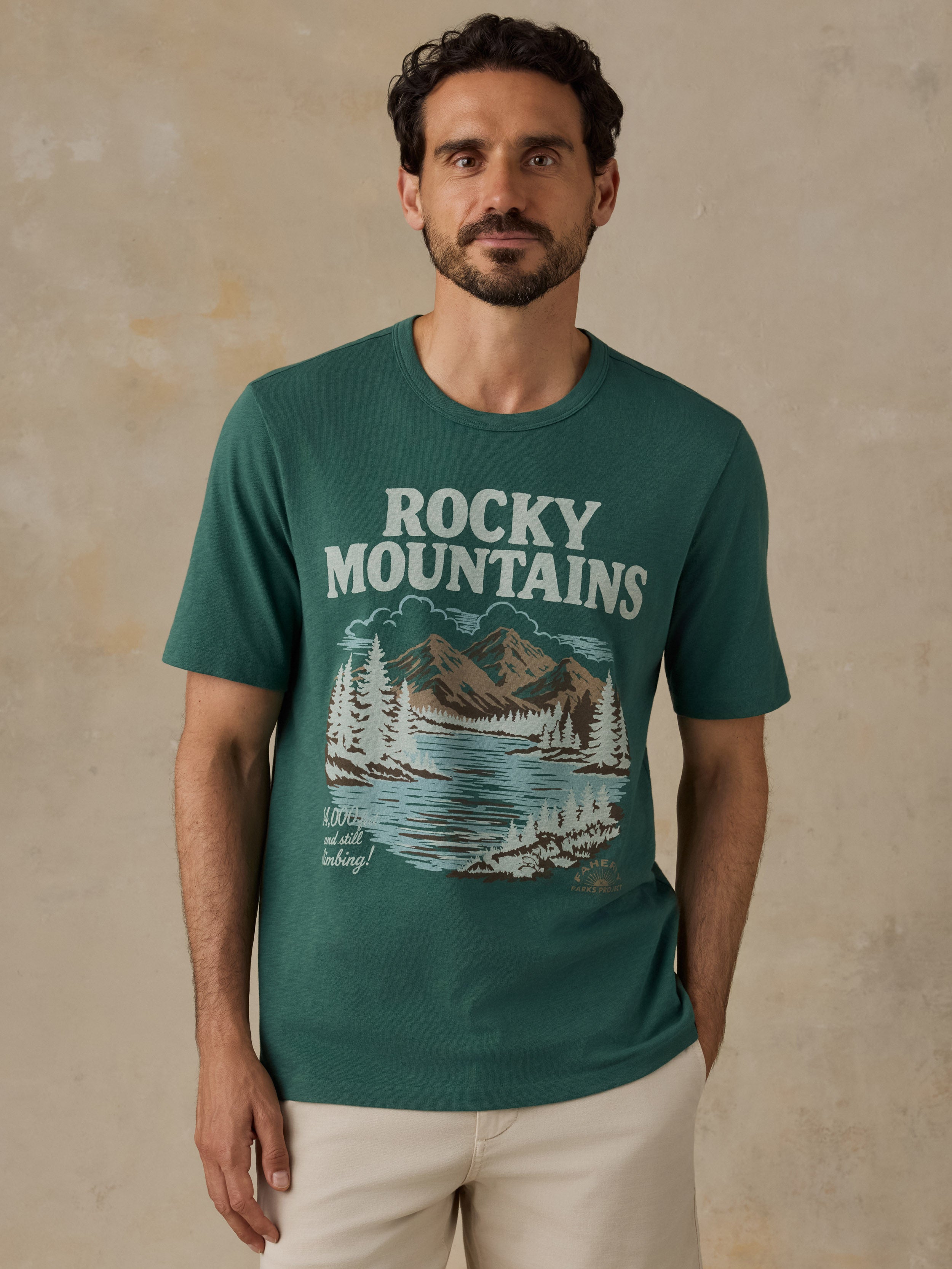 A man wearing a dark teal green t-shirt with a 'Rocky Mountains' graphic print on the front, paired with light beige shorts, standing against a textured beige wall.