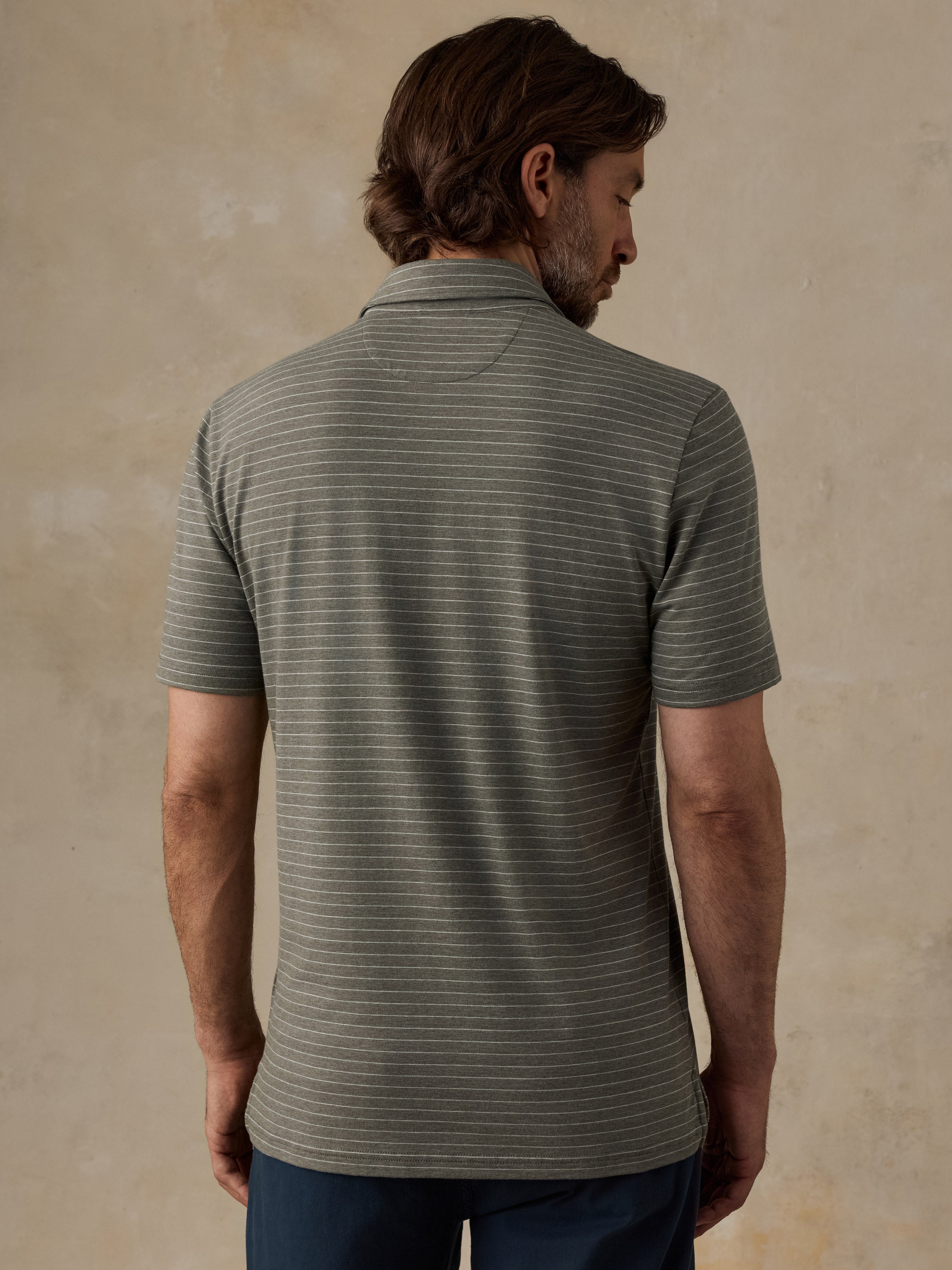 A man viewed from the back wearing a short-sleeve polo shirt with fine horizontal stripes in olive green and gray, paired with dark blue pants, set against a textured beige background.