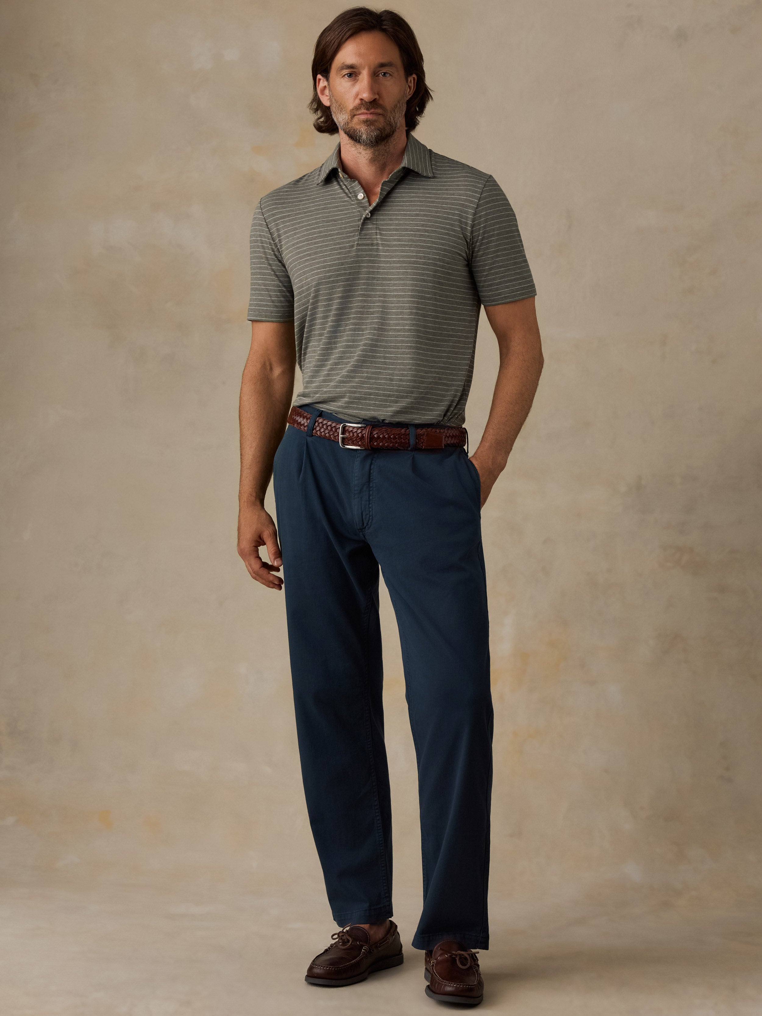 A man modeling a short-sleeve polo shirt with fine horizontal stripes in gray and olive green, paired with navy blue trousers, a brown leather belt, and brown loafers, standing against a textured beige studio backdrop.