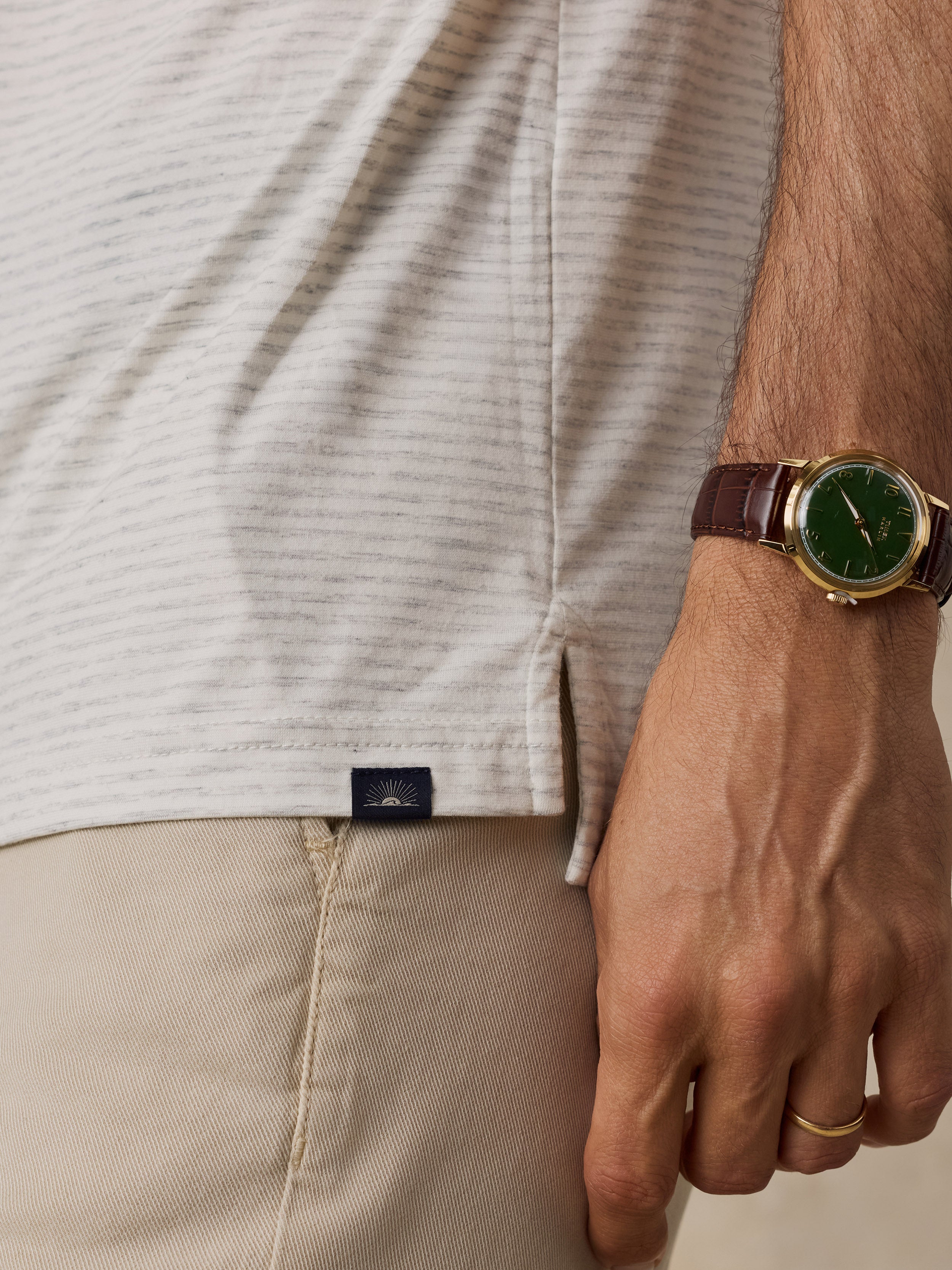 Close-up of a man wearing a light beige striped short-sleeve shirt, khaki pants, and a brown leather strap watch with a green dial.