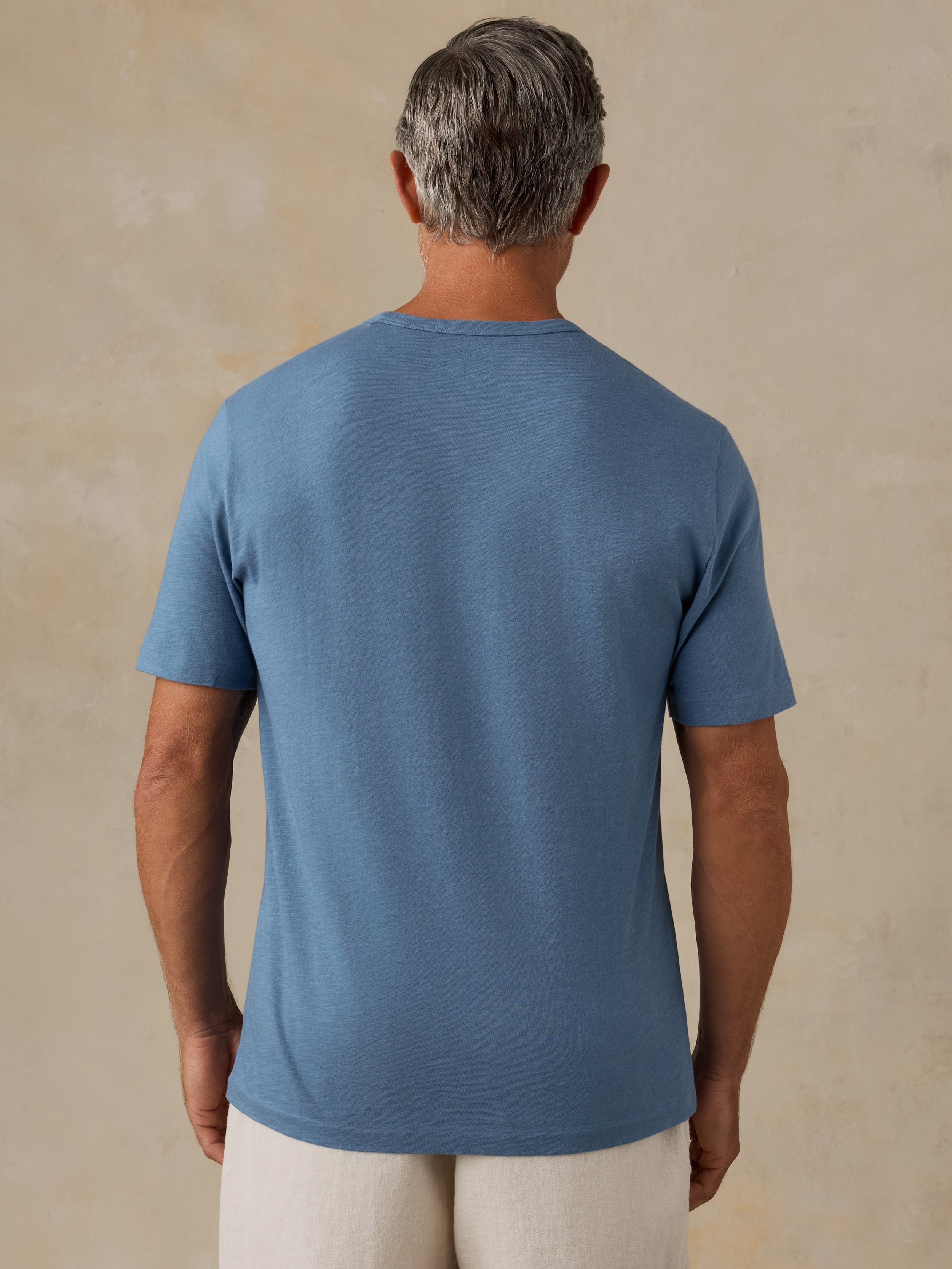A man viewed from behind wearing a heathered medium blue crewneck t-shirt and off-white pants, standing against a textured beige wall.