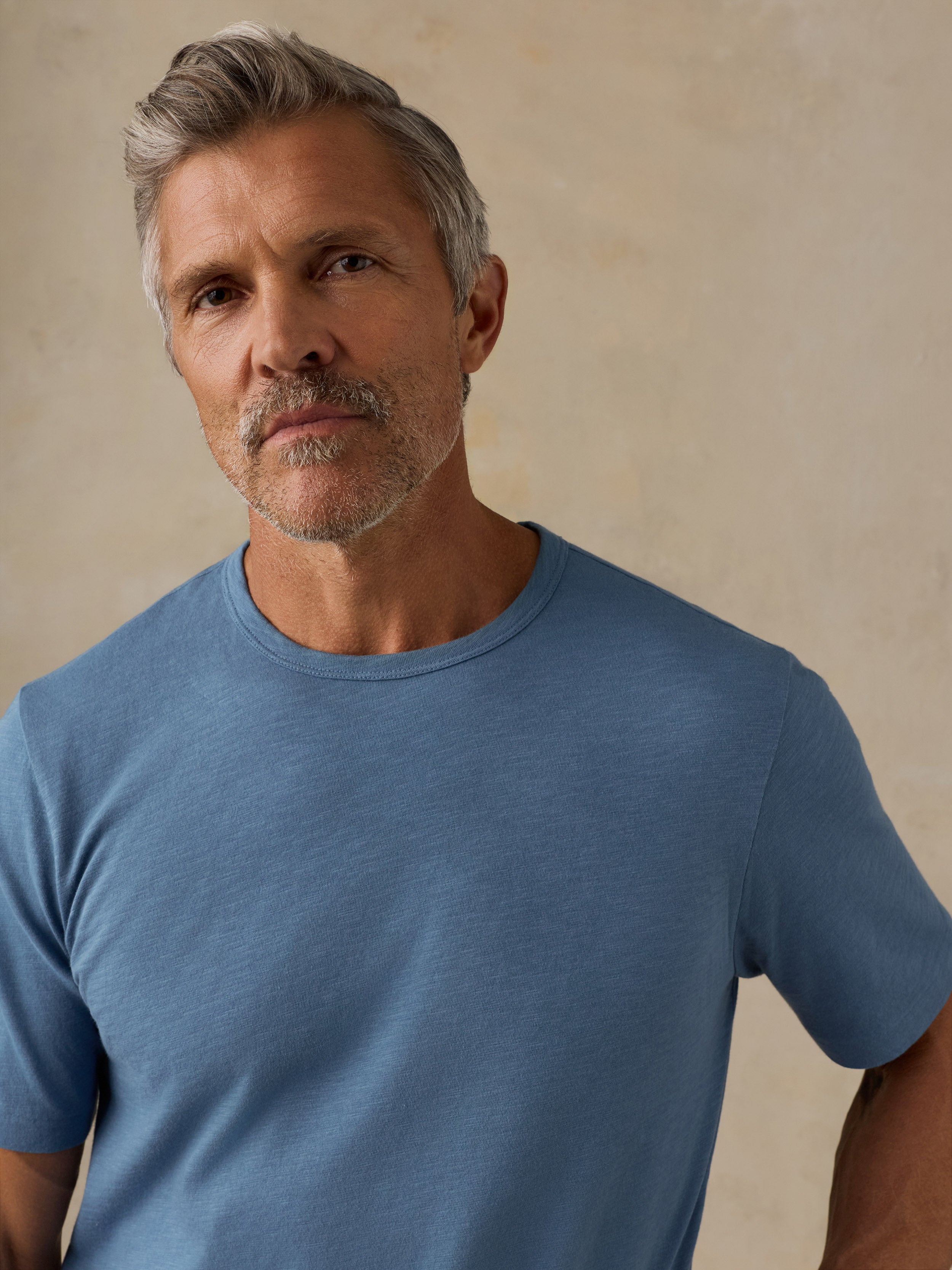 A man with gray hair and a short beard wearing a heathered blue crew-neck t-shirt, photographed against a textured beige background.