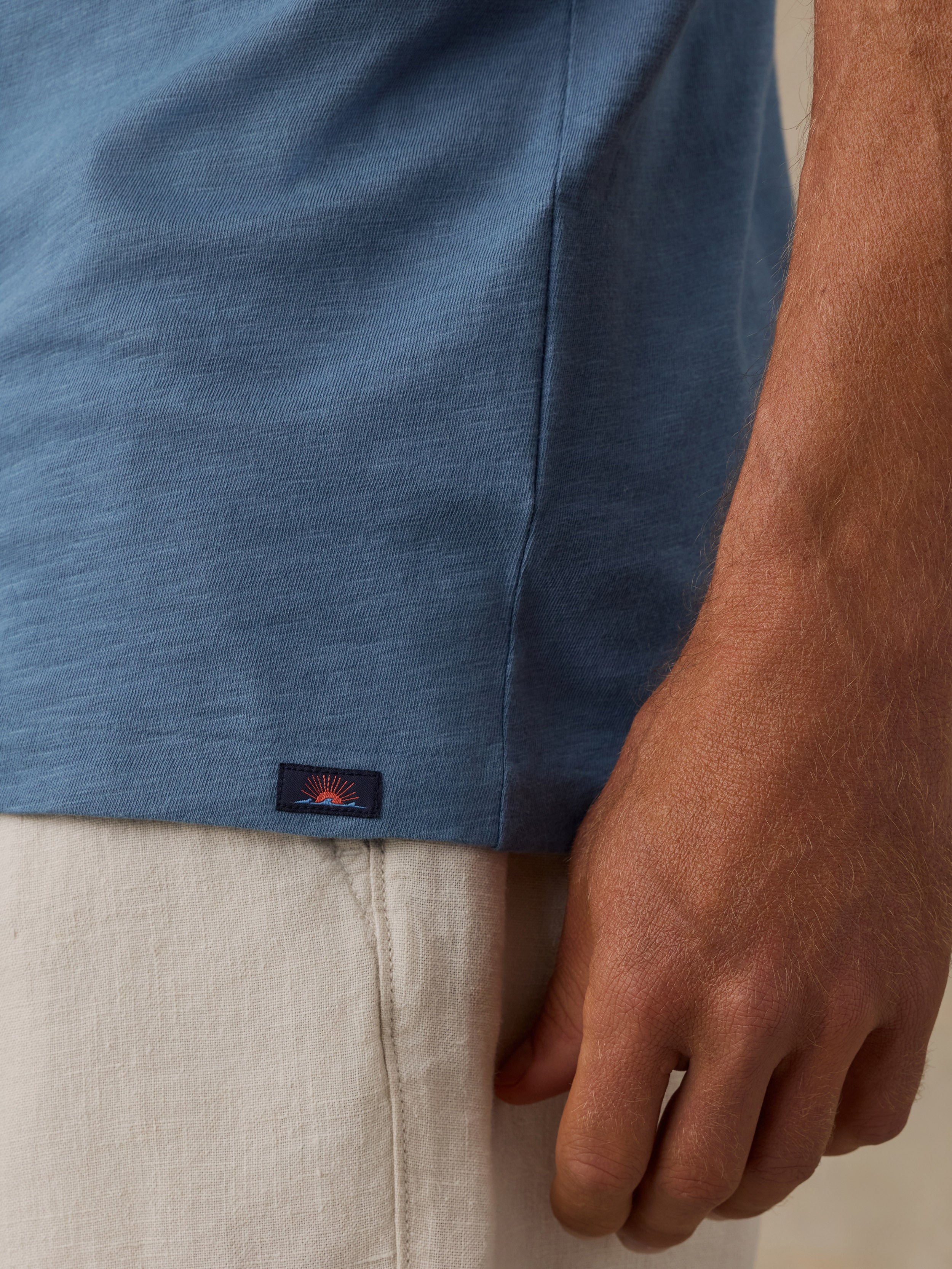 Close-up of a man wearing a heathered blue short-sleeve top and light beige pants, with a small dark Faherty brand tab near the hem.