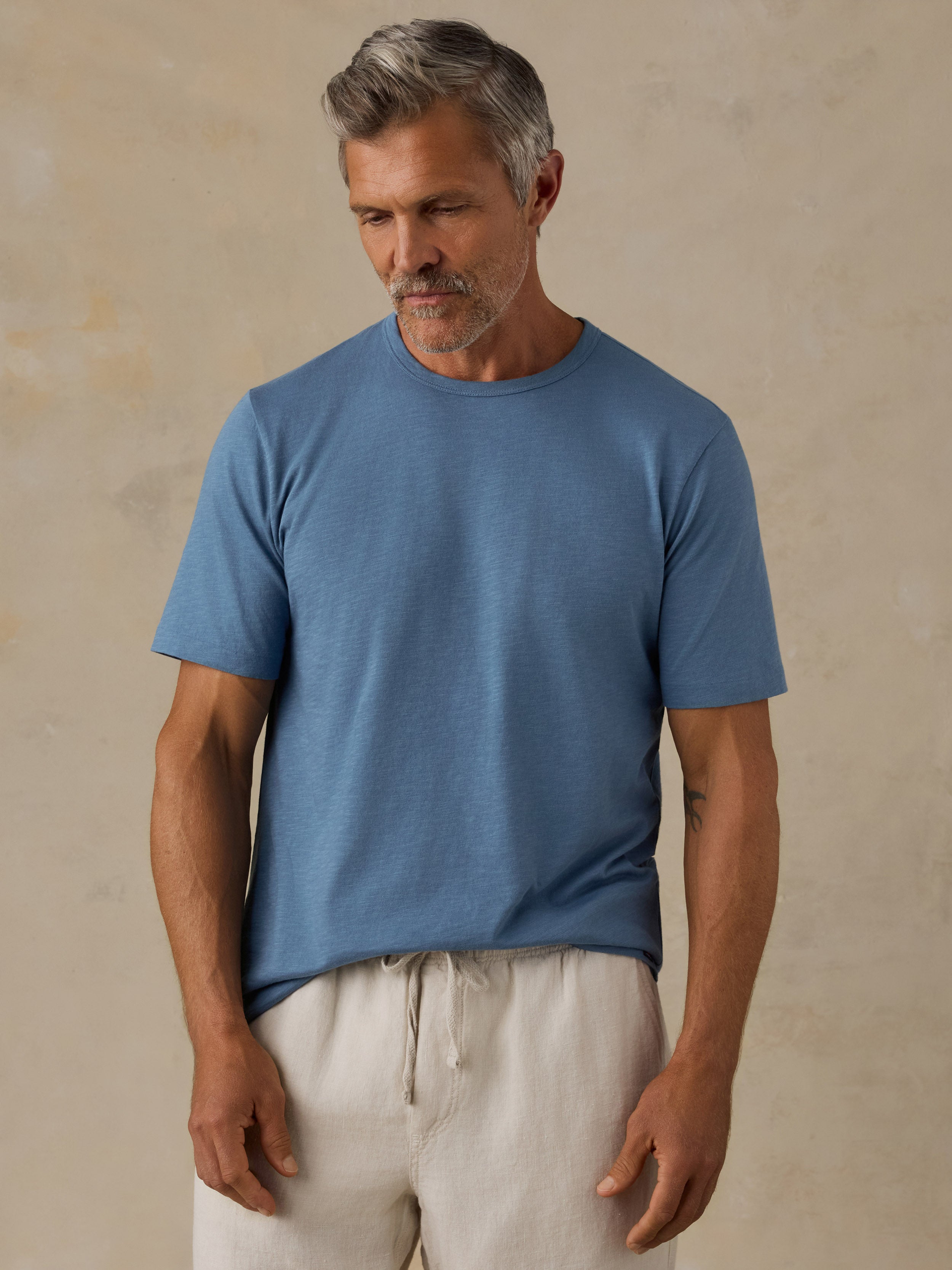 A man wearing a muted medium blue crew-neck t-shirt and off-white drawstring shorts, standing against a textured beige wall.