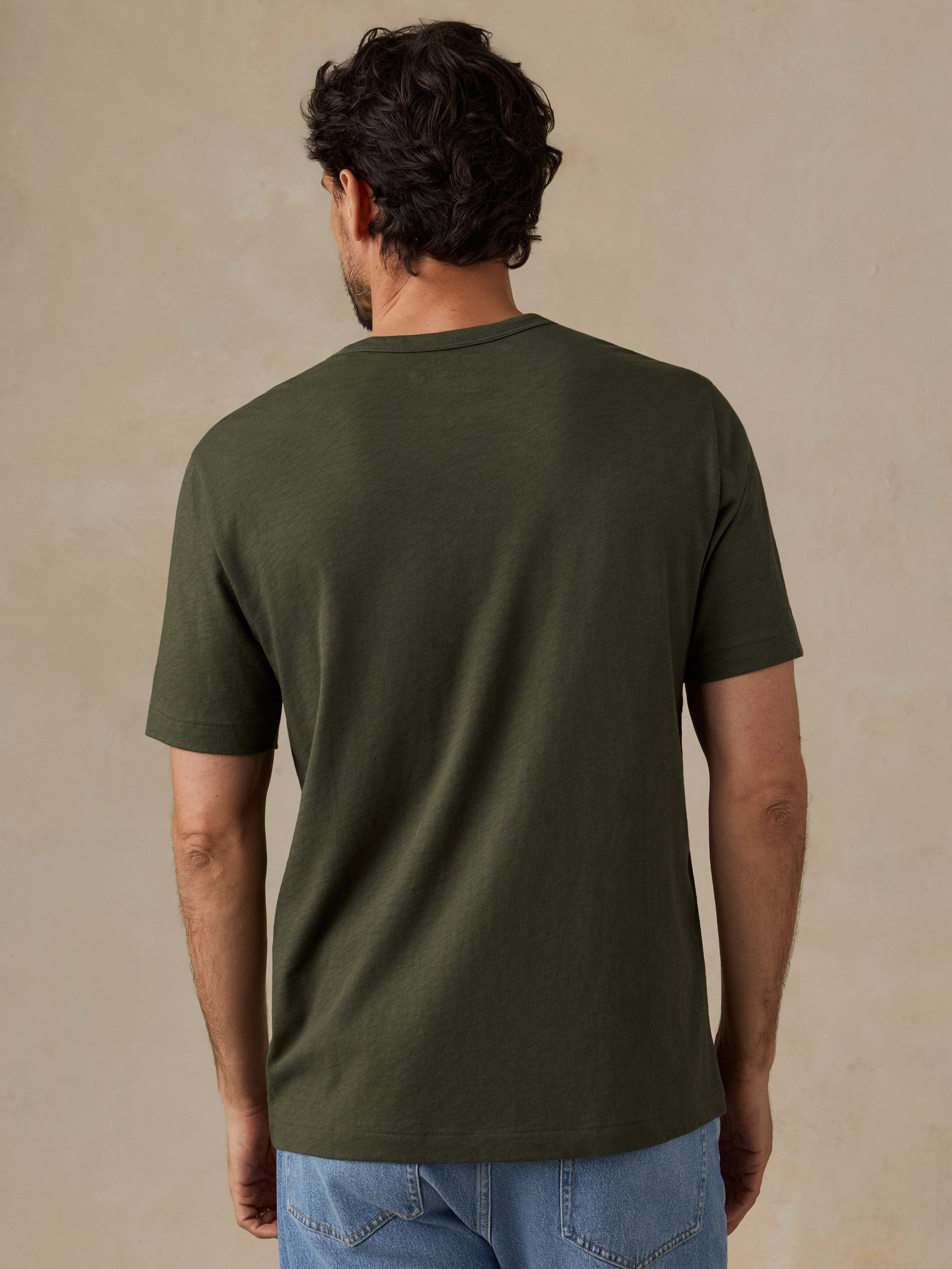 A man viewed from behind wearing a dark olive green short-sleeve t-shirt and light blue jeans, standing against a textured beige studio background.