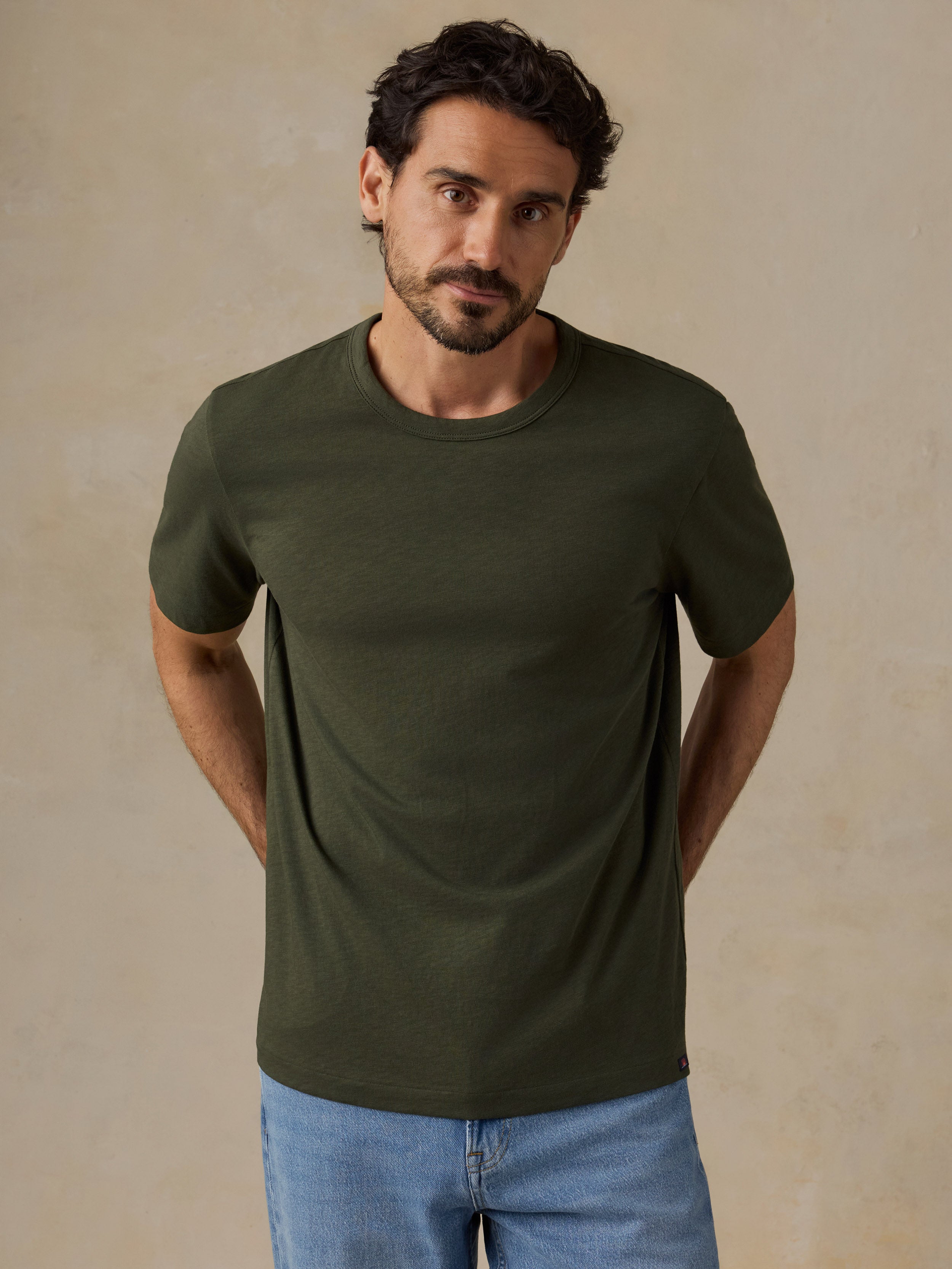 A man wearing a dark olive green short-sleeve crewneck t-shirt and light blue jeans, standing against a textured beige wall.
