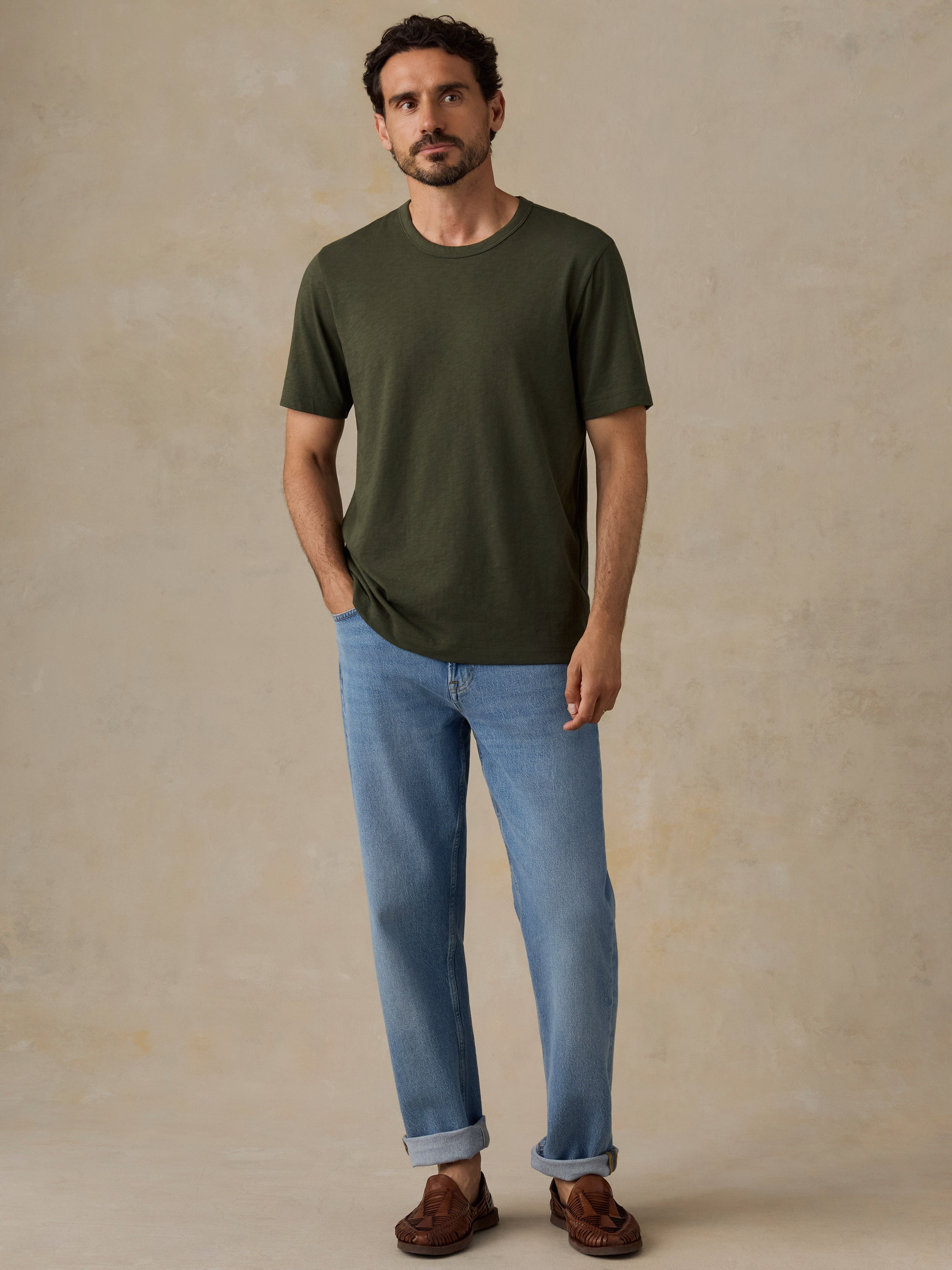 A man wearing a dark olive green crew-neck t-shirt, light blue jeans, and brown leather loafers, standing against a textured beige studio backdrop.