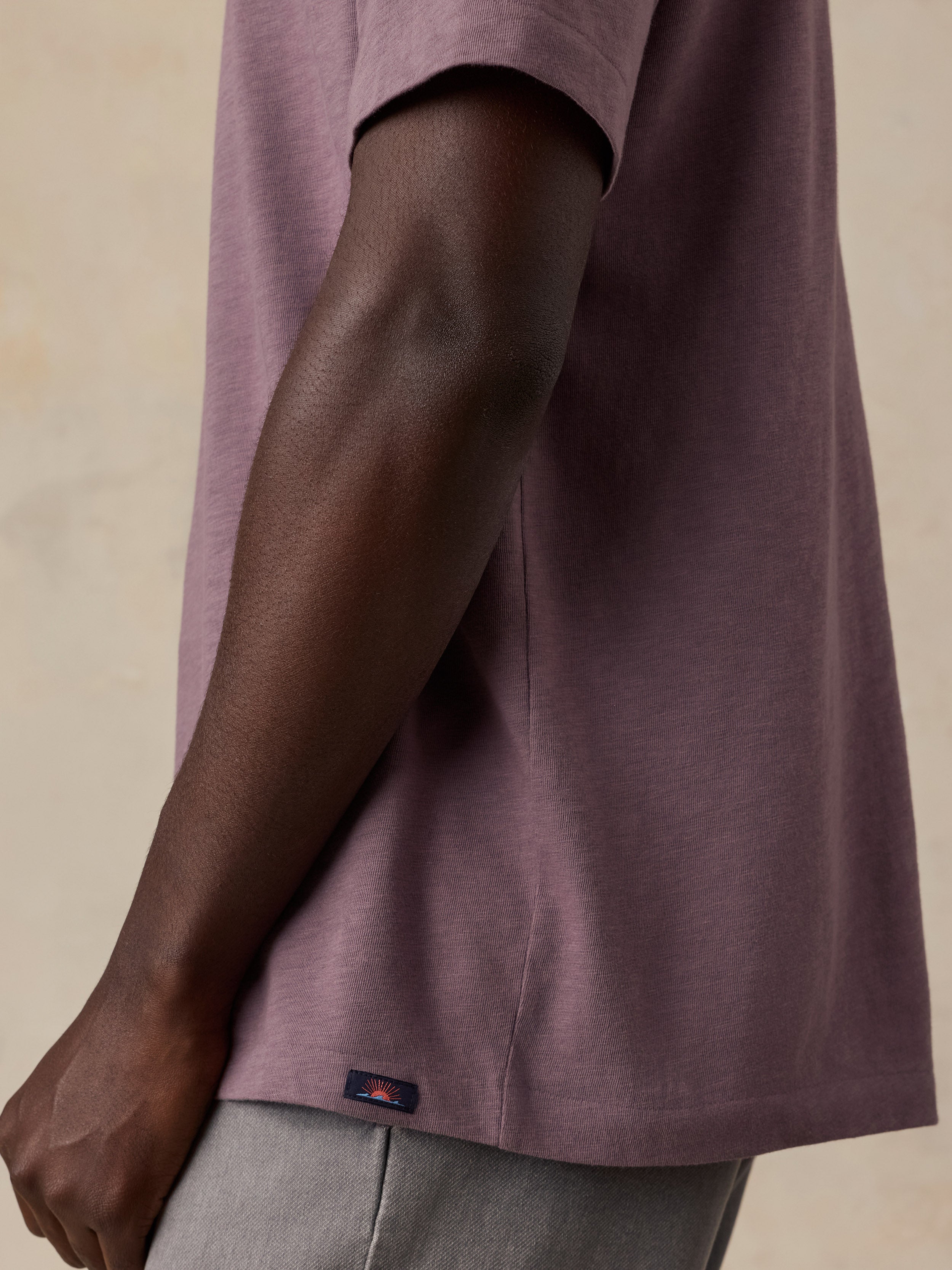 Close-up side view of a man wearing a muted mauve-purple short-sleeve T-shirt and light gray pants, with a small dark Faherty brand tab near the hem.