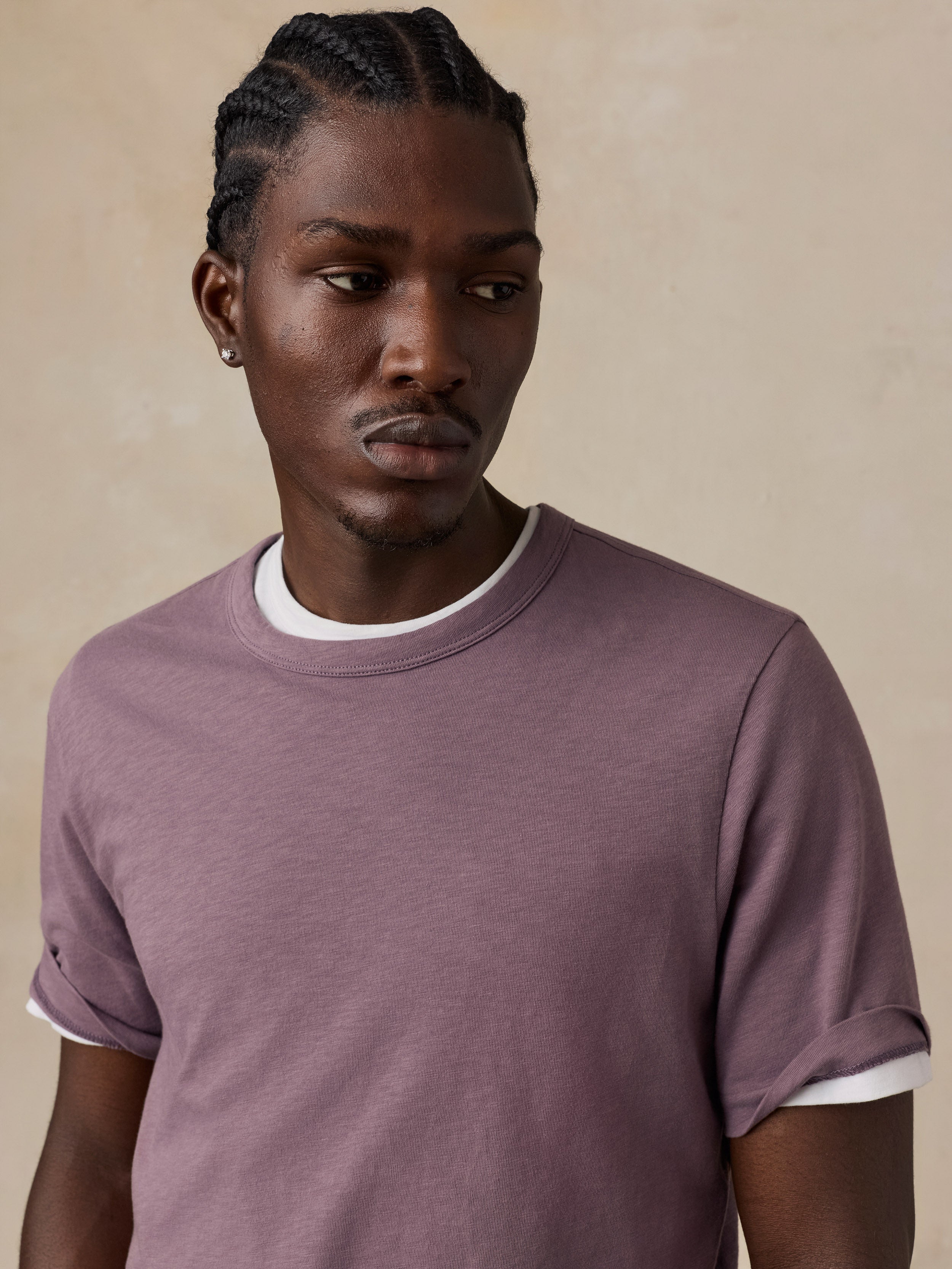 A man wearing a dusty mauve short-sleeve t-shirt set against a textured beige background.