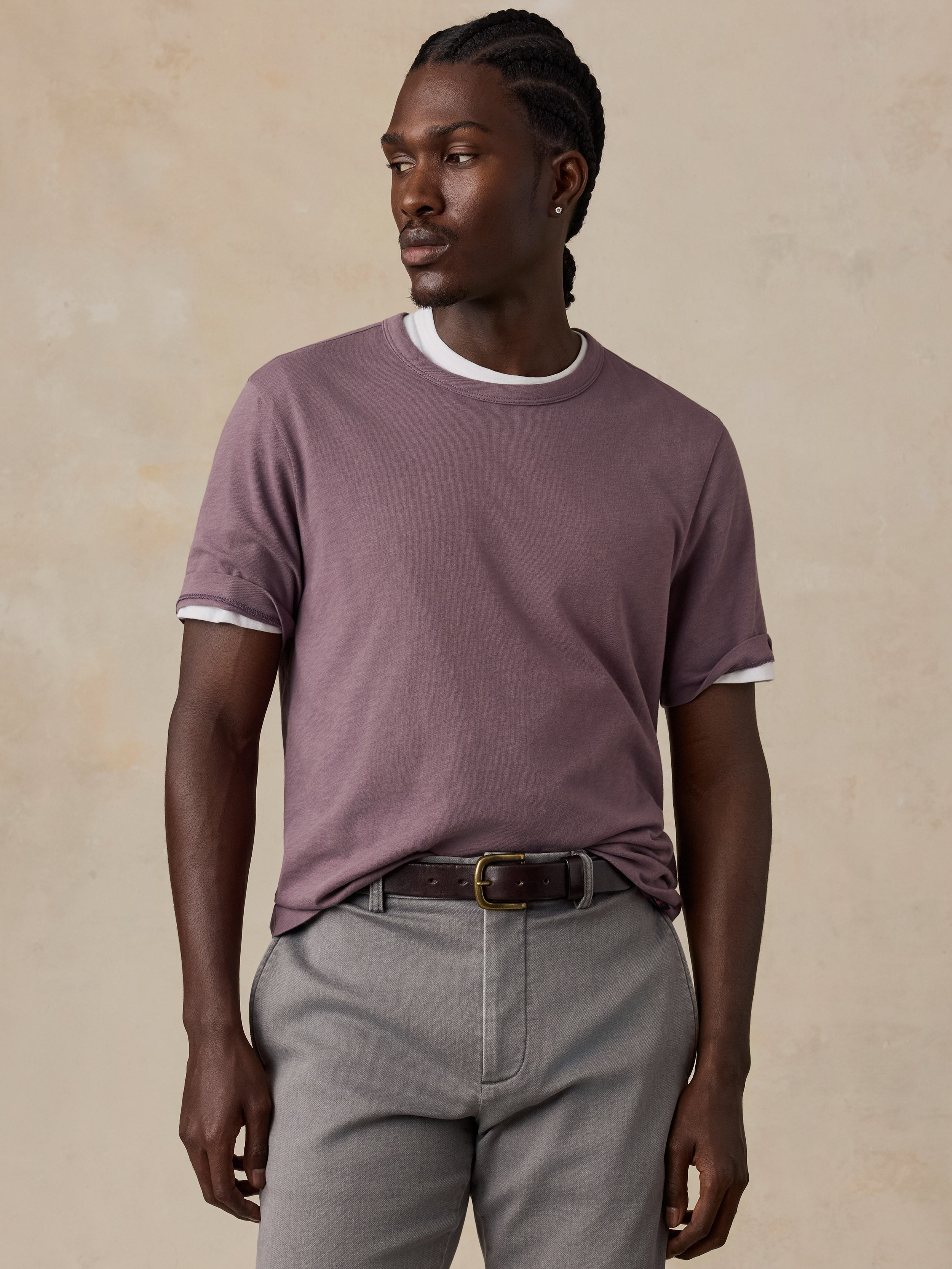 A man wearing a mauve t-shirt, paired with gray trousers and a brown belt, standing against a textured beige background.