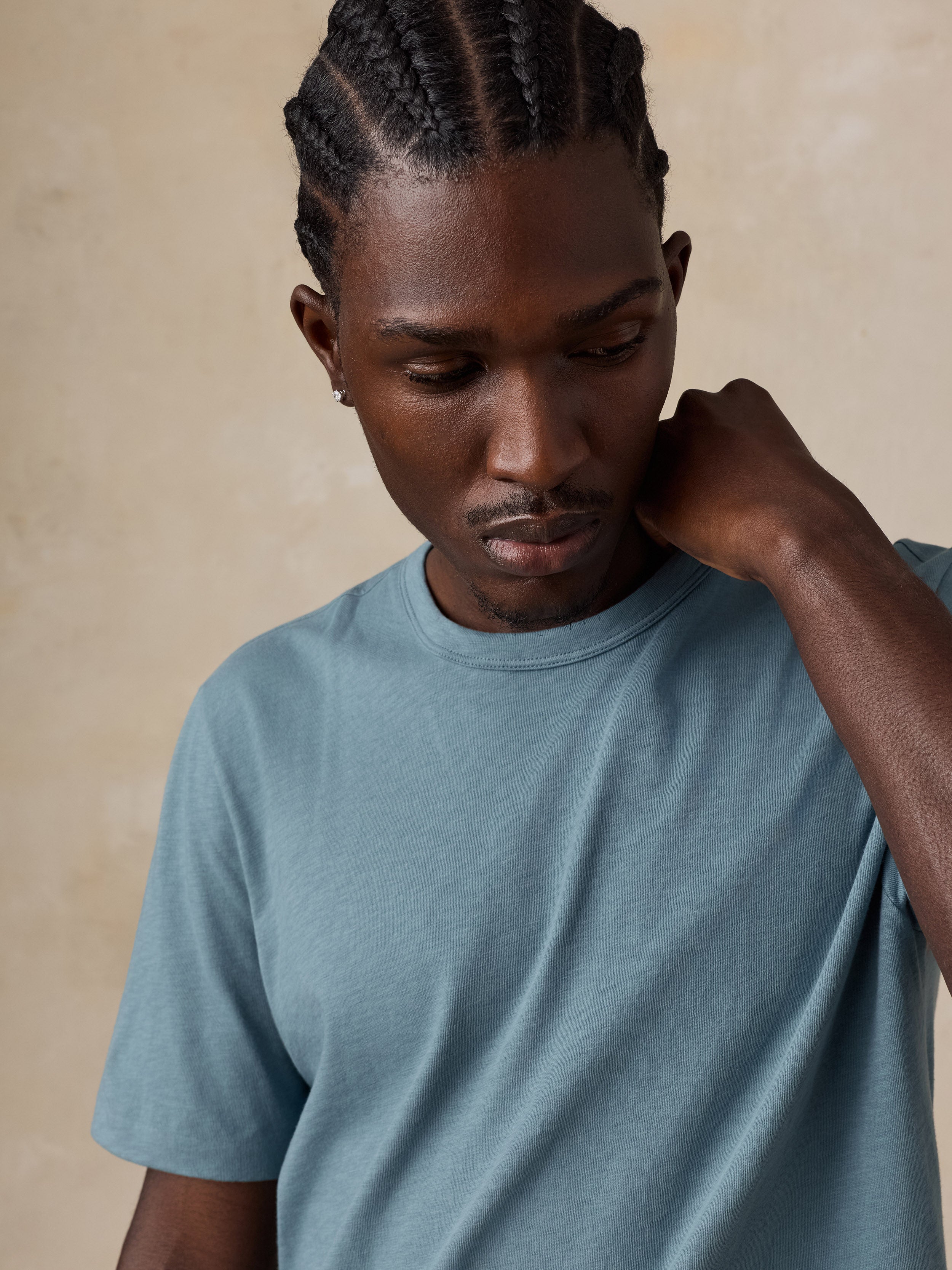A man with braided hair wearing a muted blue-gray crewneck t-shirt, posing with his hand on his neck against a textured beige background.