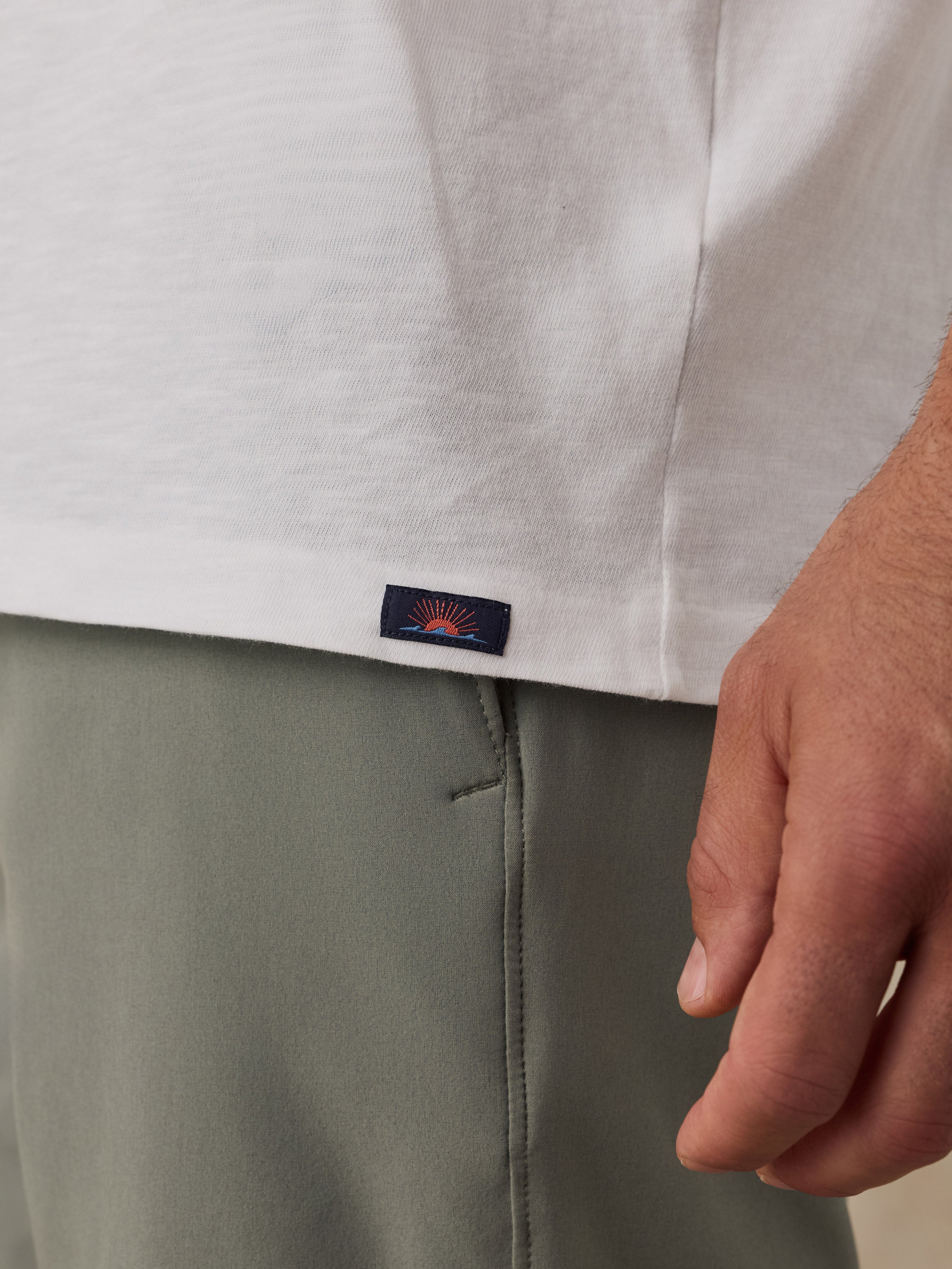 Close-up of a man wearing a white t-shirt and olive green shorts, showing a small rectangular Faherty brand patch on the hem of the shirt.