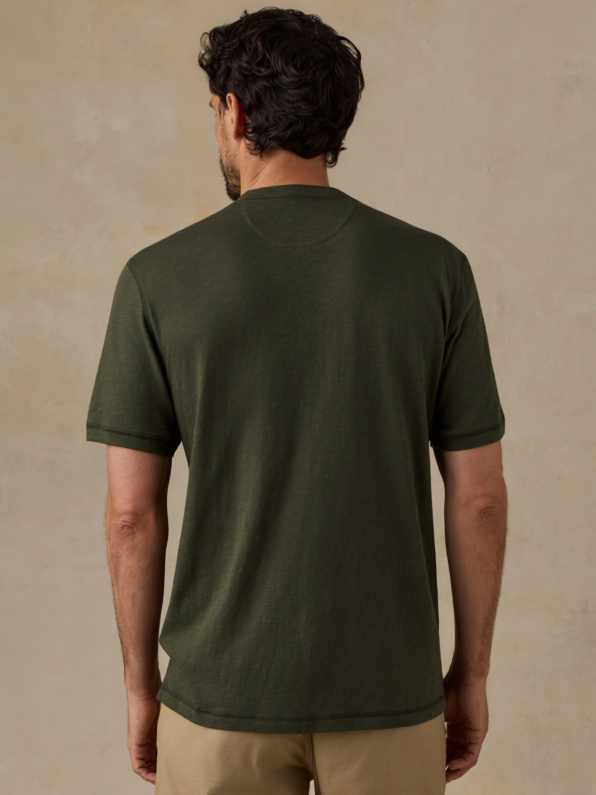 A man viewed from behind wearing an olive green short-sleeve crewneck t-shirt and khaki shorts, standing against a textured beige wall.