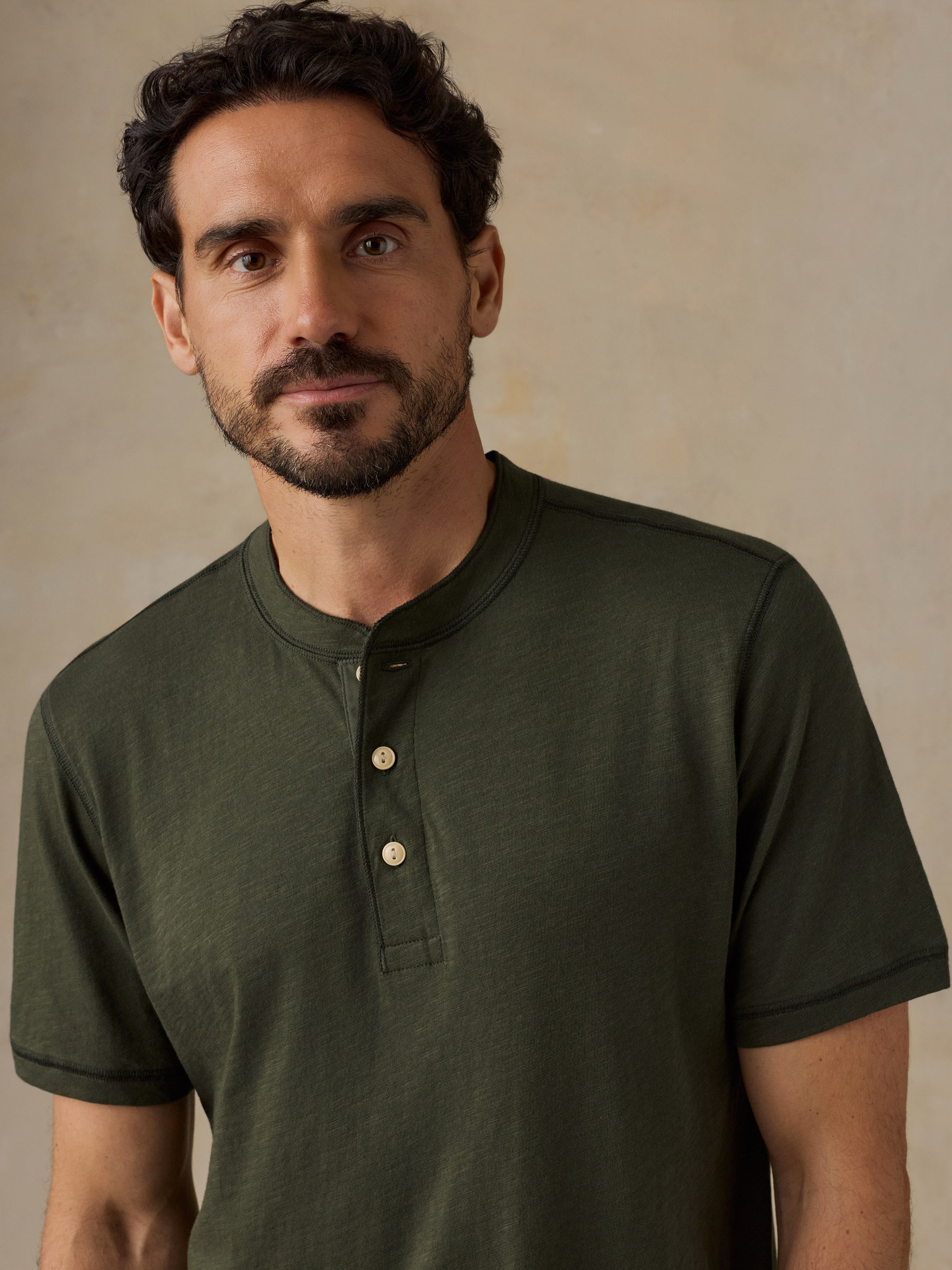 A man wearing a dark olive green short-sleeve henley shirt with a three-button placket, photographed against a neutral textured beige background.