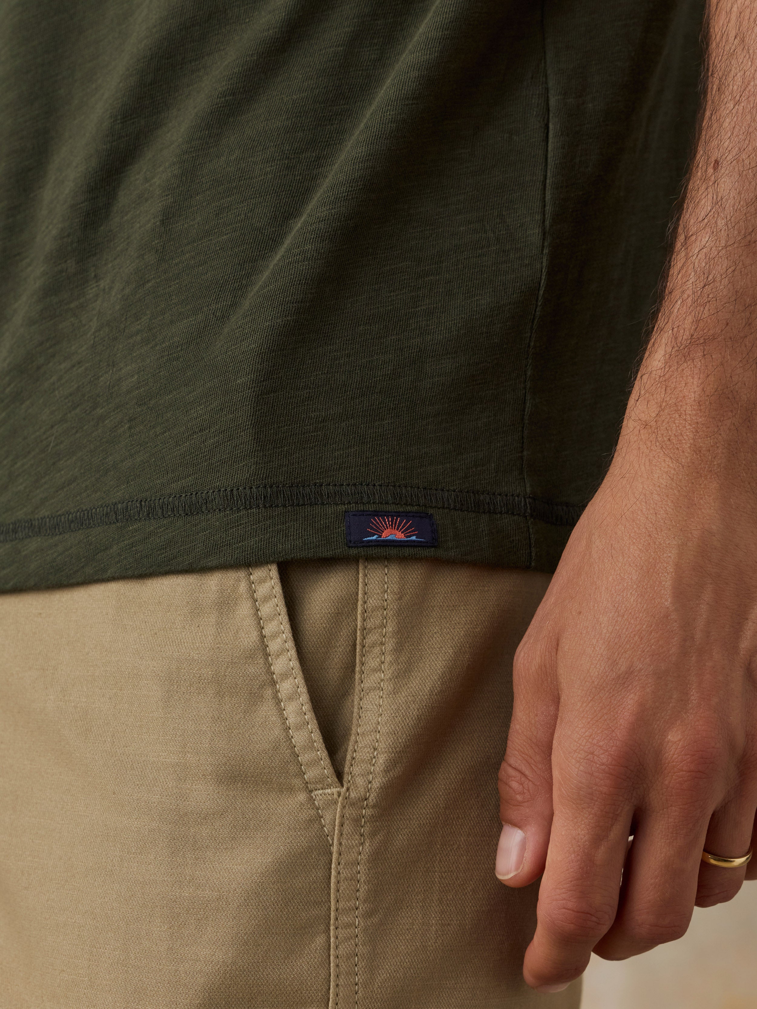 Close-up of a man wearing an olive green short-sleeve t-shirt and khaki shorts, showing a small Faherty brand logo patch on the hem.