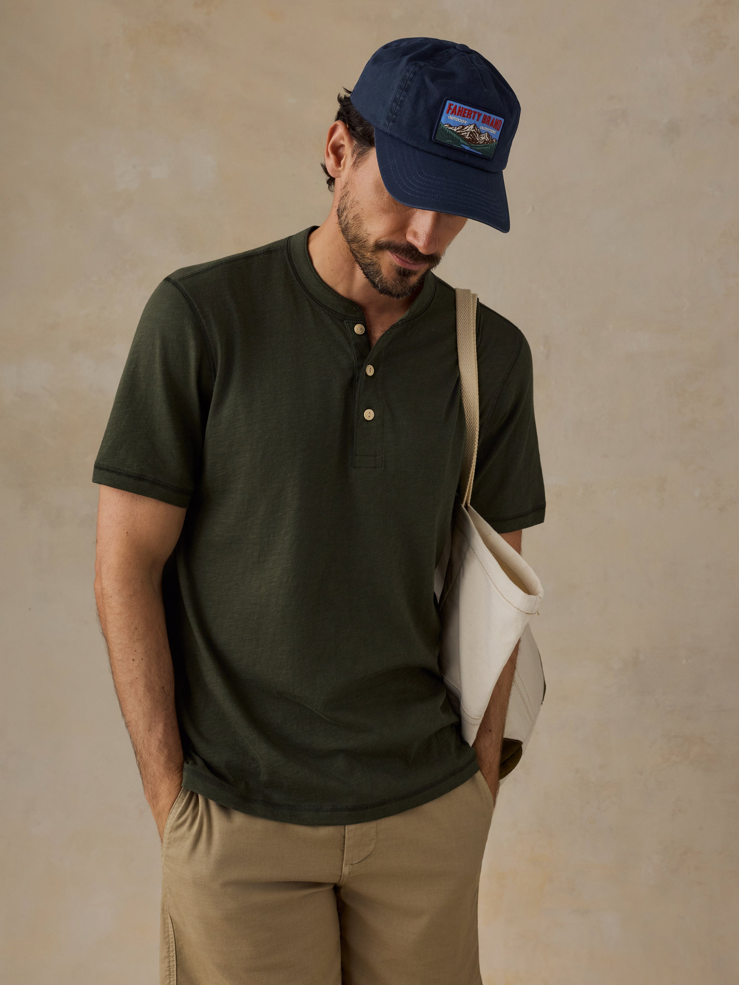 A man wearing a dark olive green short-sleeve henley shirt, khaki shorts, a navy blue cap, and carrying a cream canvas shoulder bag, set against a textured beige studio backdrop.
