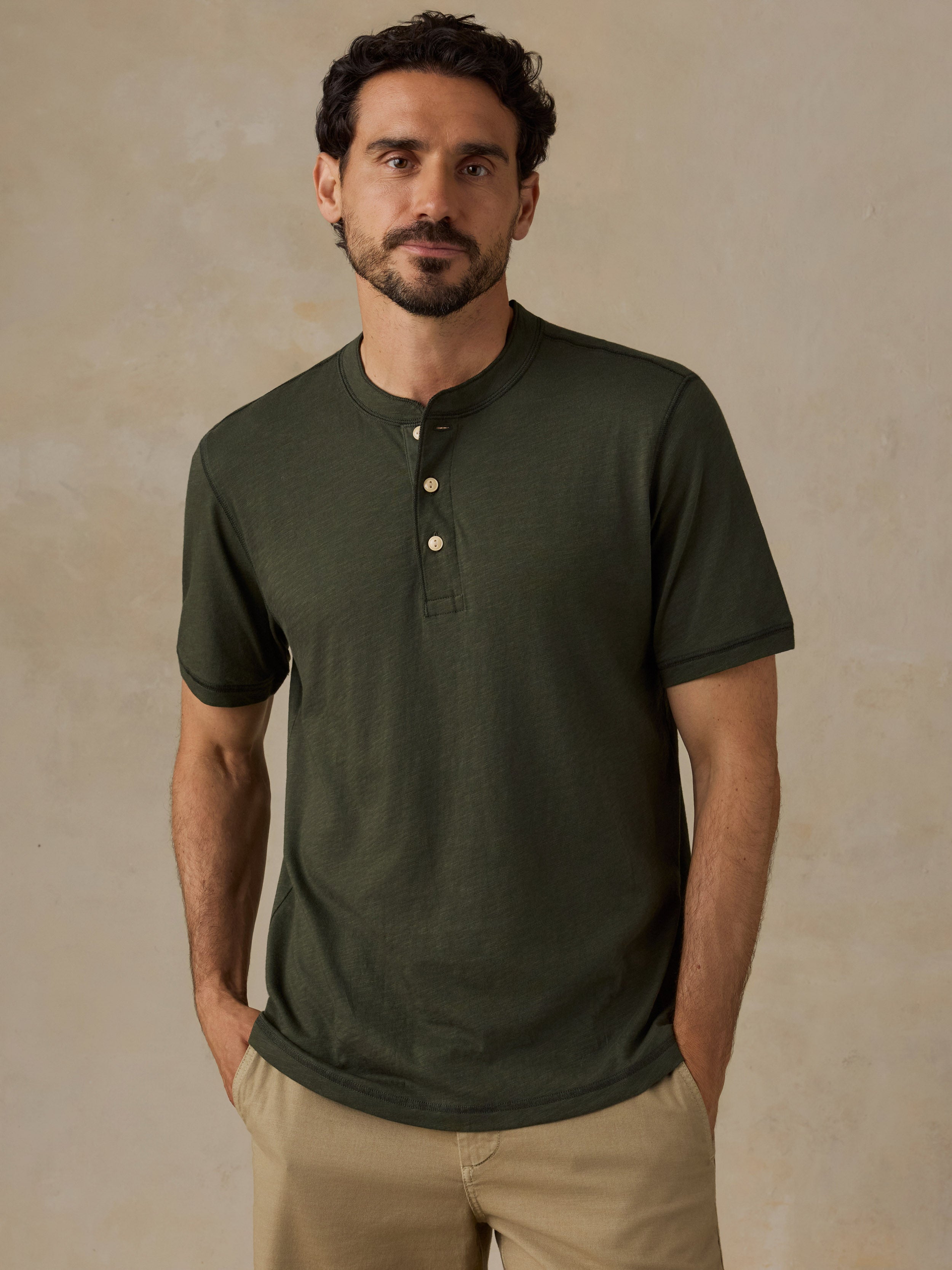 A man wearing a dark olive green short-sleeve henley shirt with three buttons, paired with khaki shorts, standing against a textured beige studio background.