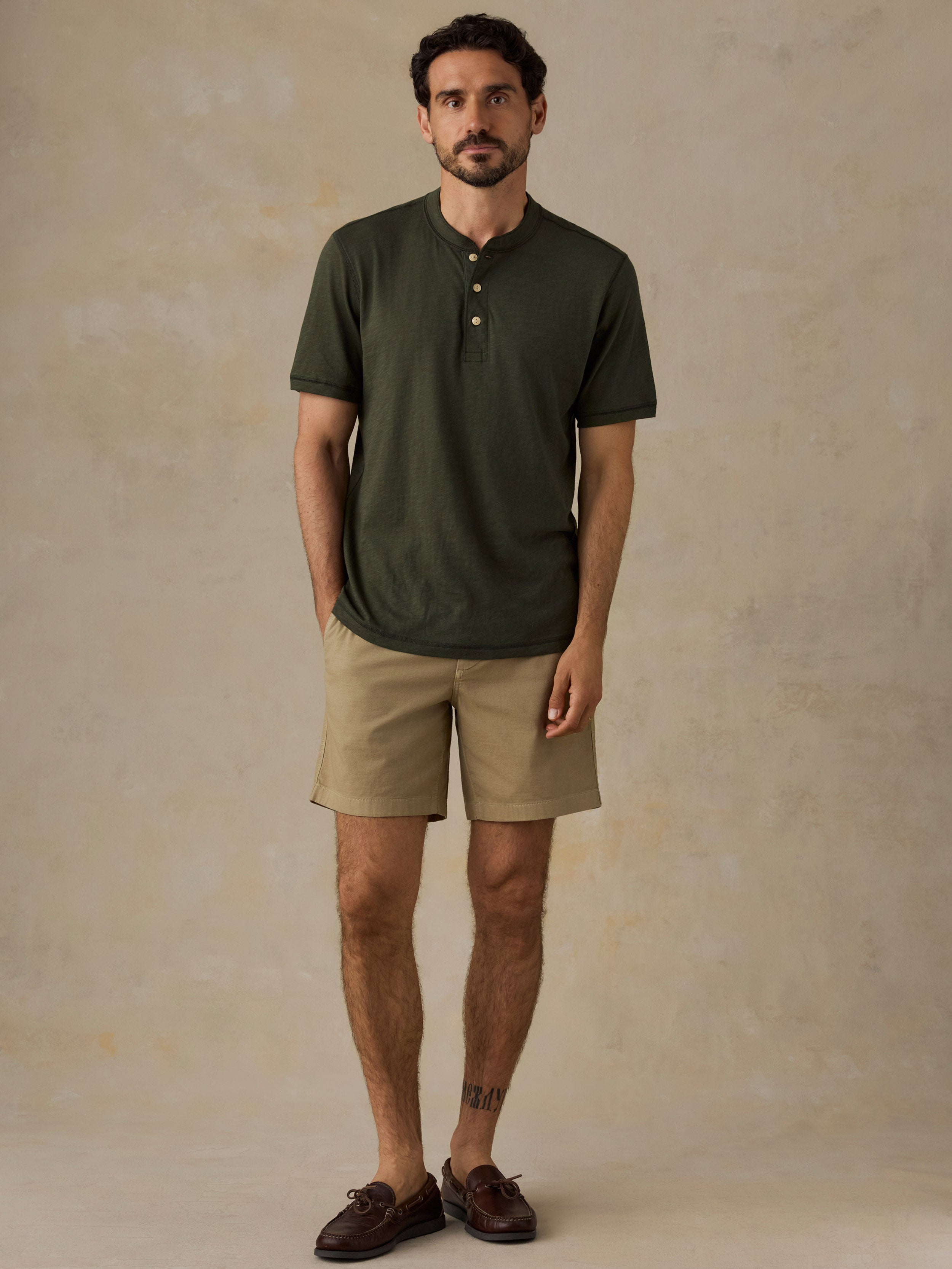 A man wearing an olive green short-sleeve henley shirt, beige shorts, and dark brown loafers, standing against a textured beige studio backdrop.