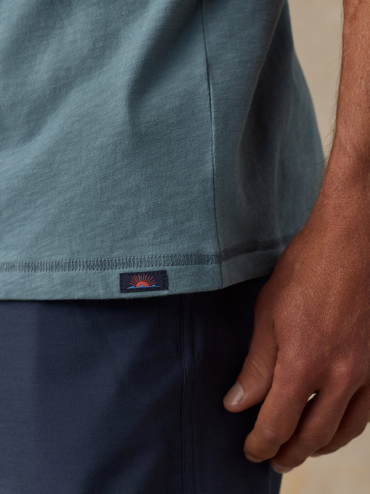 Close-up of a man wearing a muted blue-gray short-sleeve t-shirt and dark navy shorts, with a small embroidered logo patch visible near the hem of the shirt., image 5 of 6