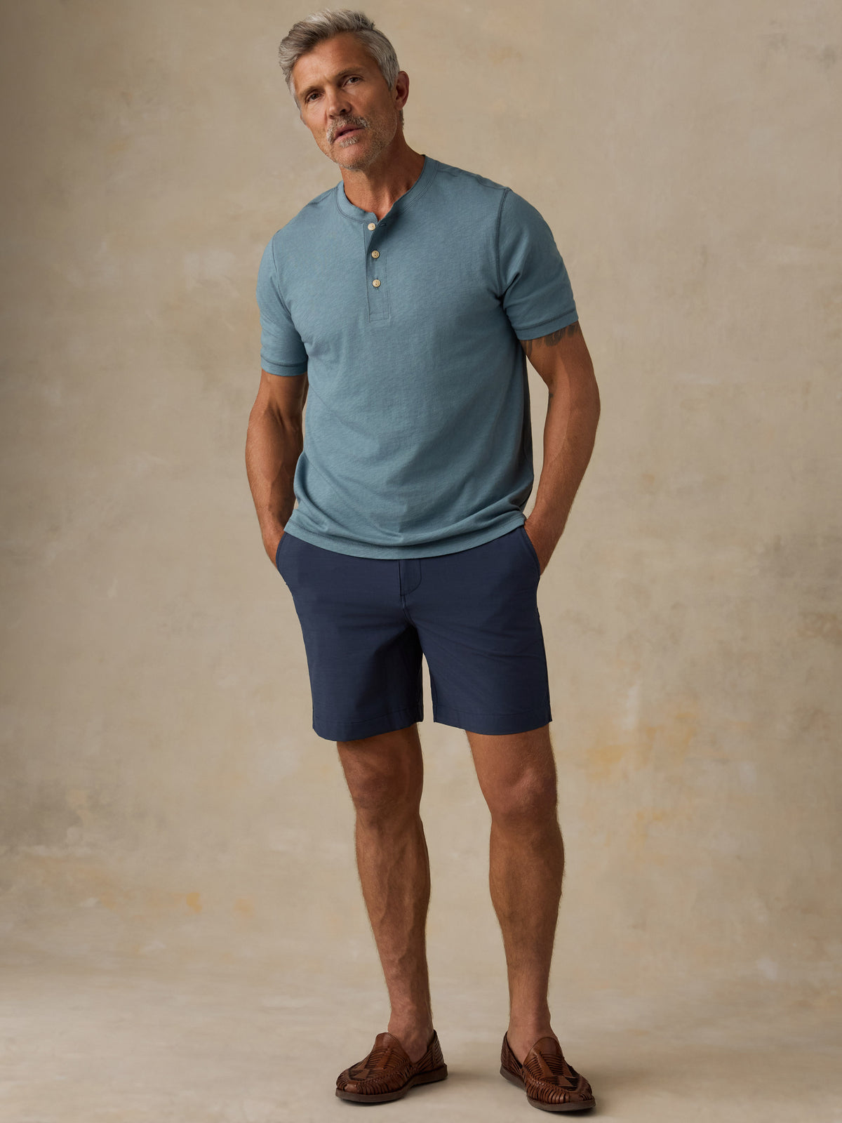 A man wearing a dusty blue short-sleeve henley shirt and navy blue shorts, standing against a textured beige studio backdrop., image 6 of 6