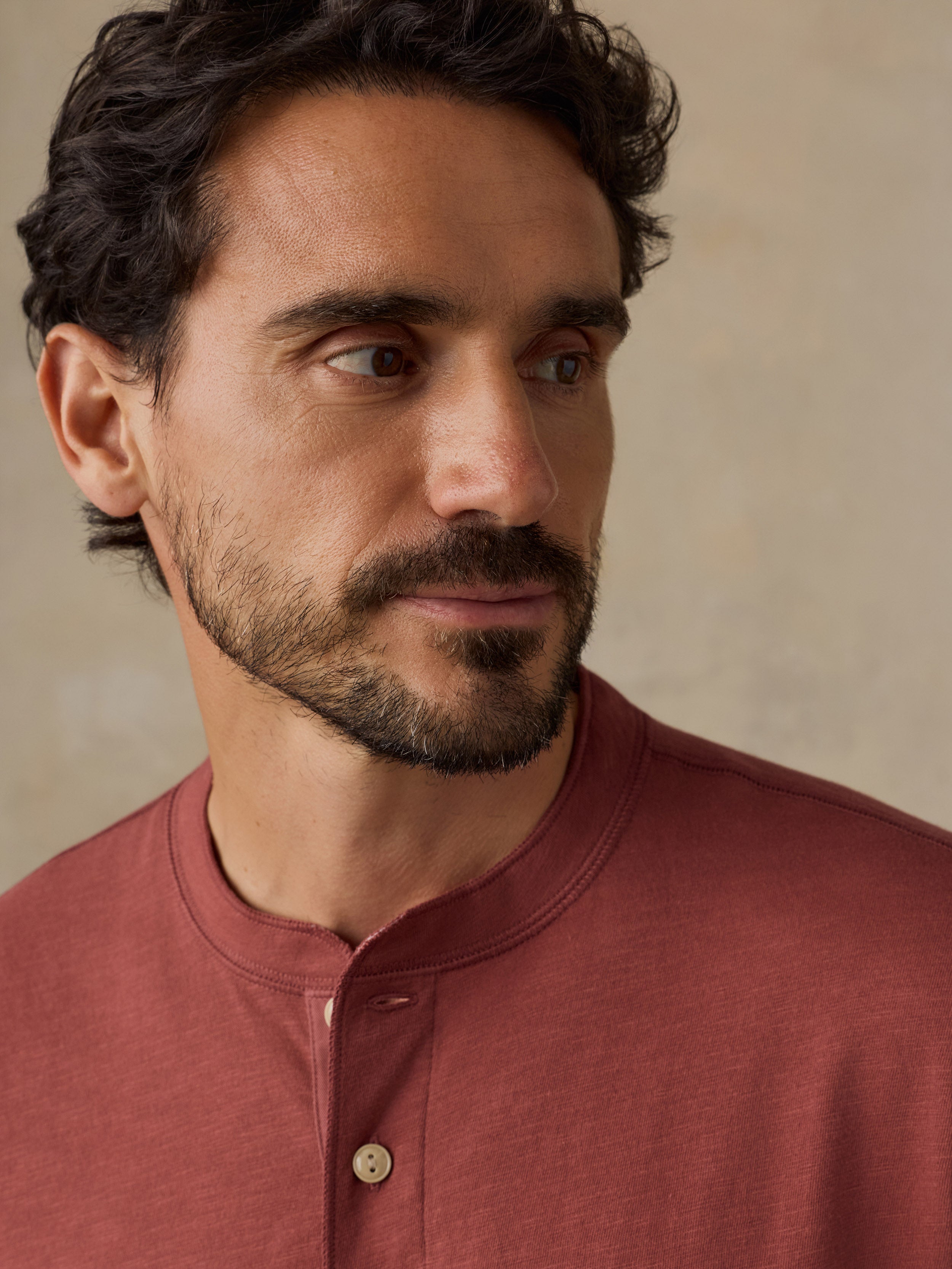 A close-up portrait of a man wearing a muted dusty rose henley shirt against a soft beige textured background.