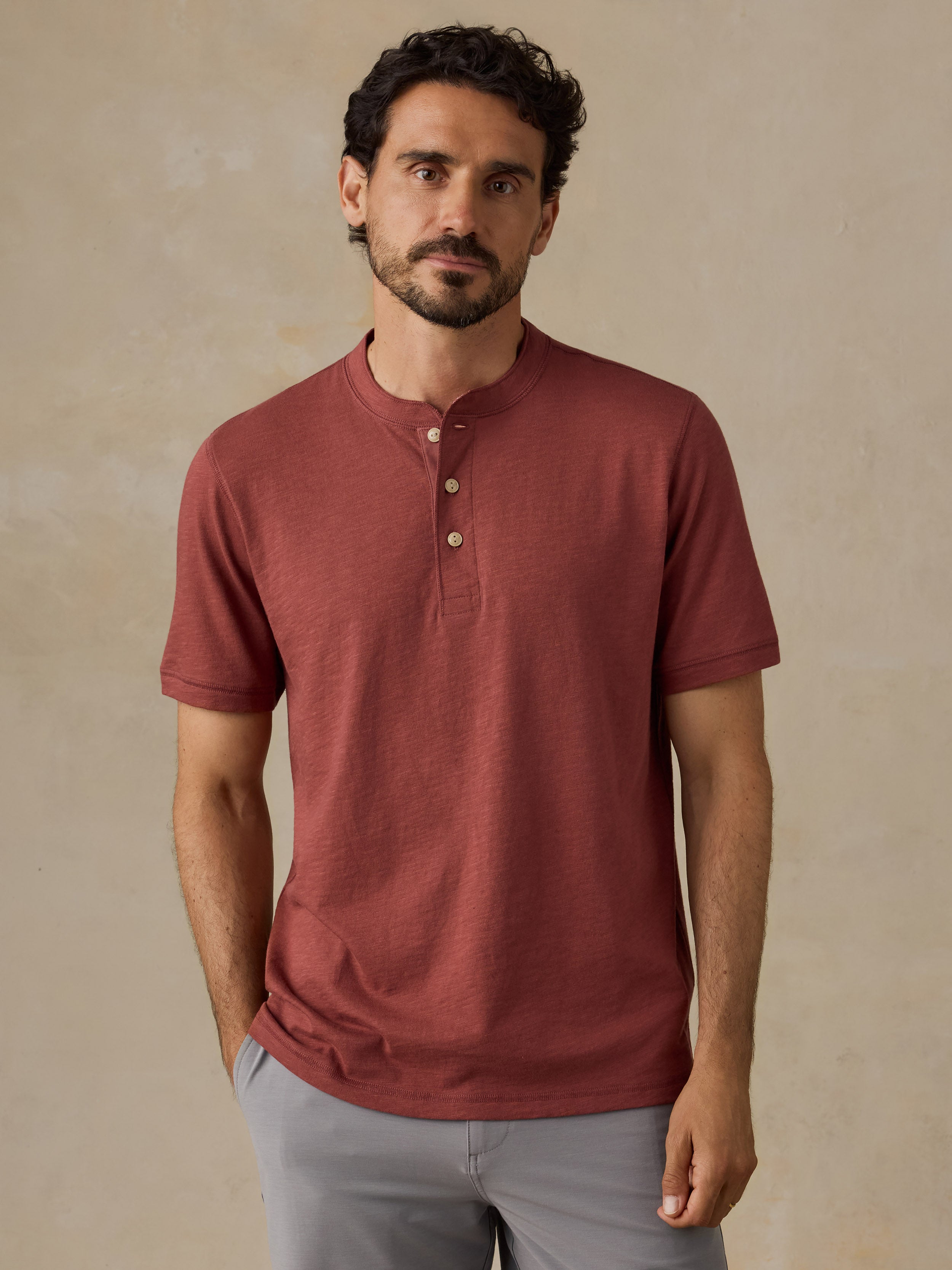 A man wearing a short-sleeve henley shirt in a muted brick red color, paired with light gray shorts, standing against a textured beige background.