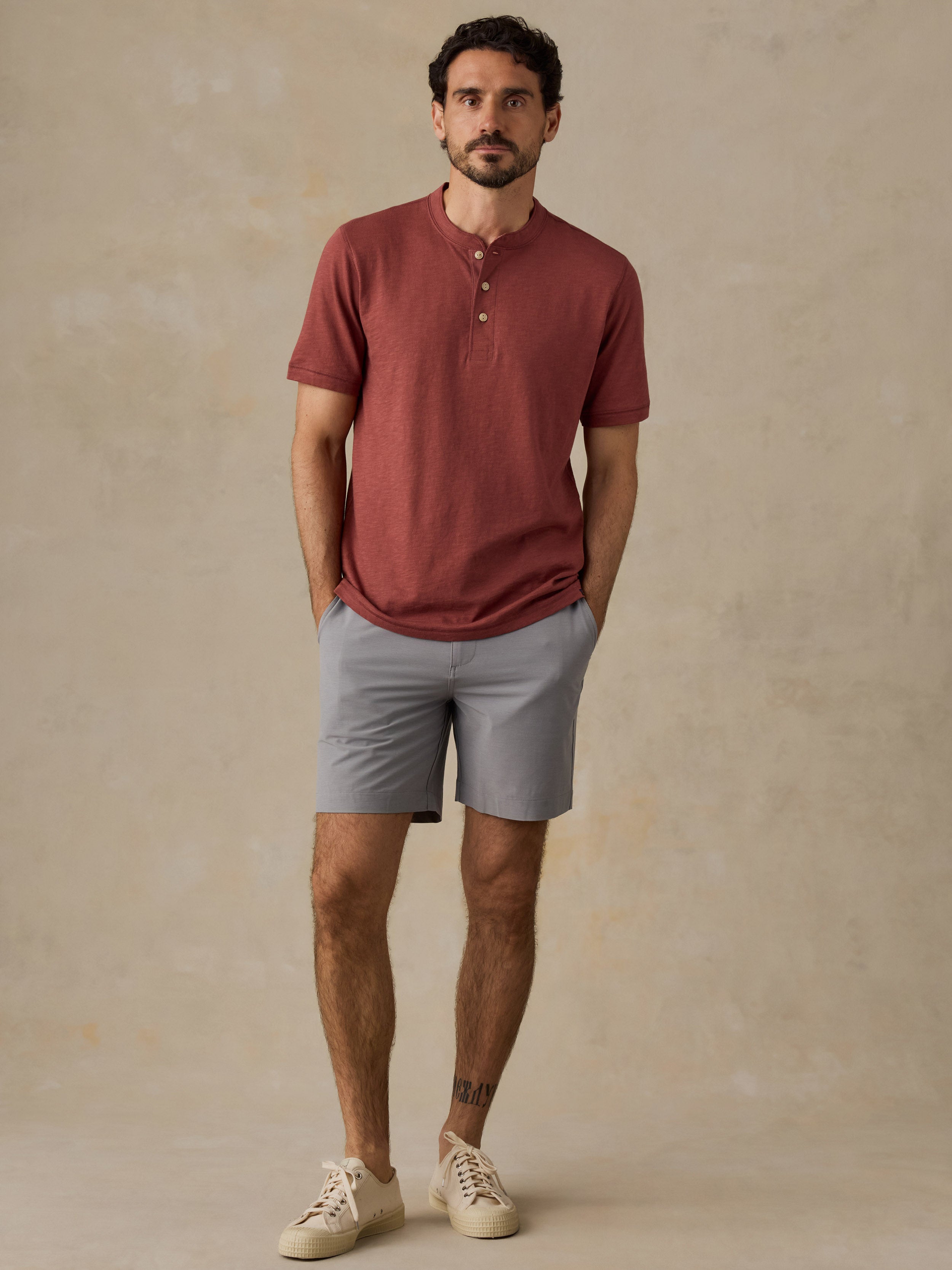 A man wearing a muted red henley t-shirt, light gray shorts, and beige sneakers, standing against a textured beige studio backdrop.