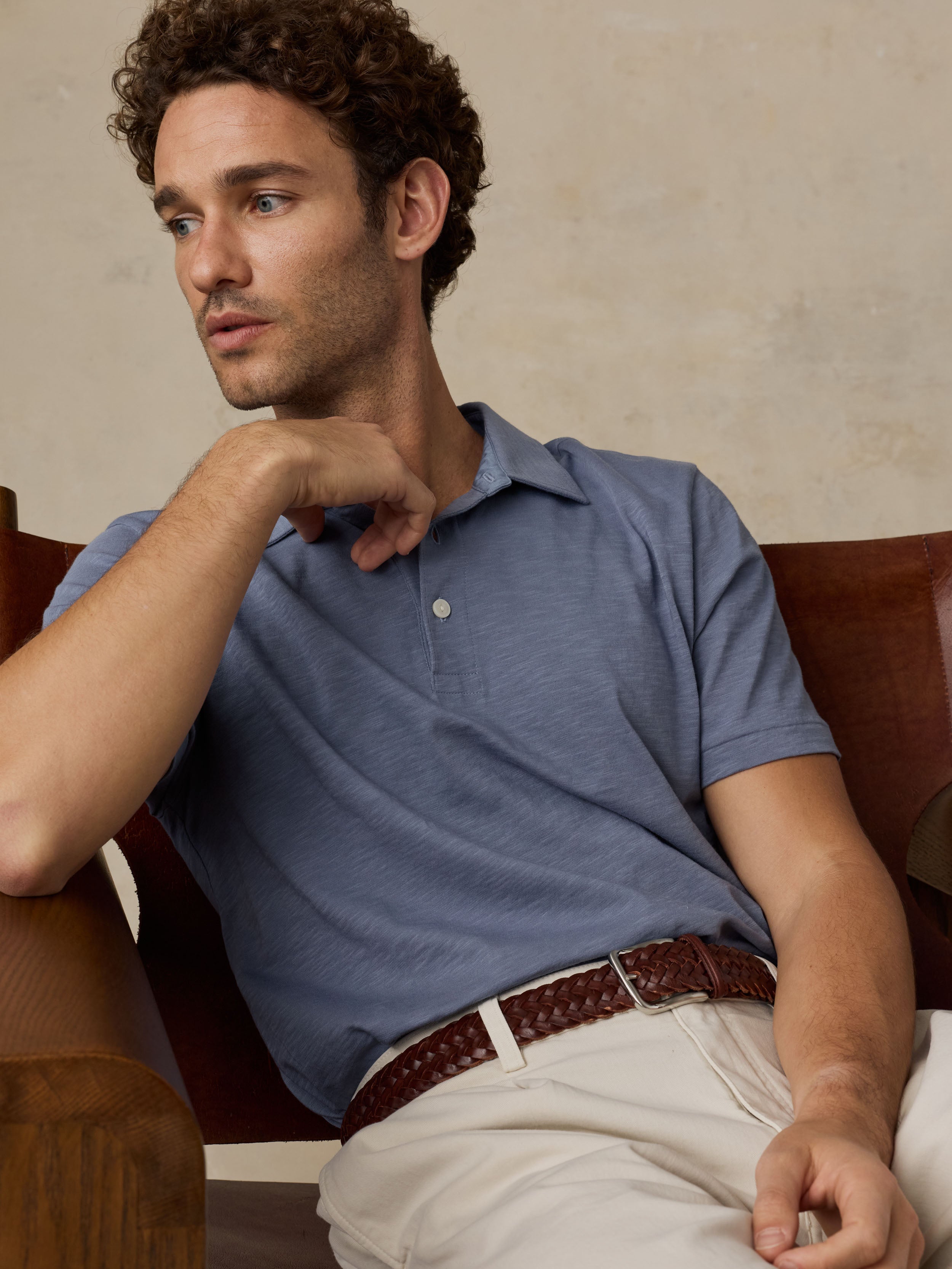 Man sitting on a wooden chair wearing a blue polo shirt and beige pants against a beige background