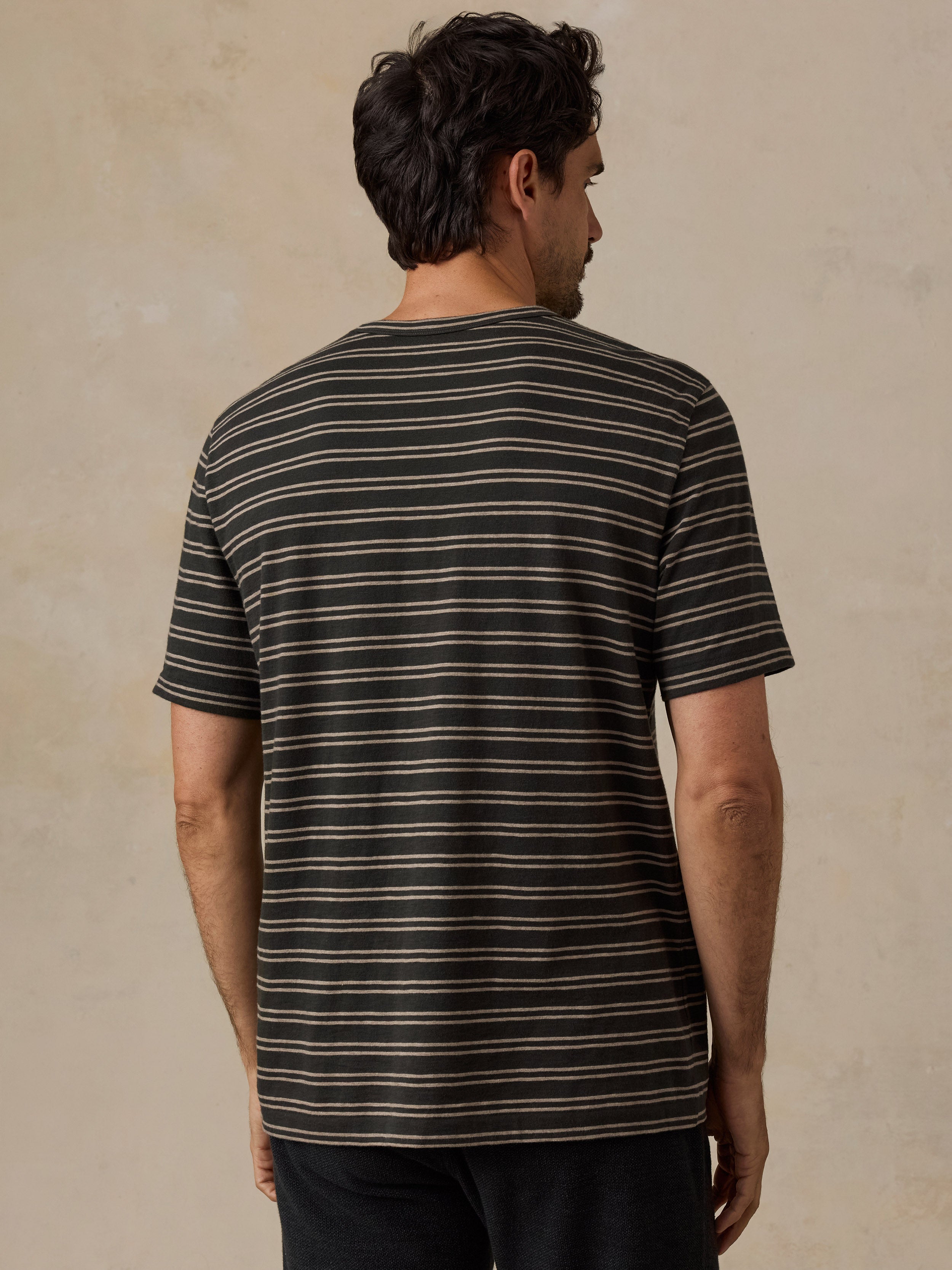 A man viewed from behind wearing a short-sleeve crewneck t-shirt with thin horizontal stripes in black and beige, set against a textured beige wall.