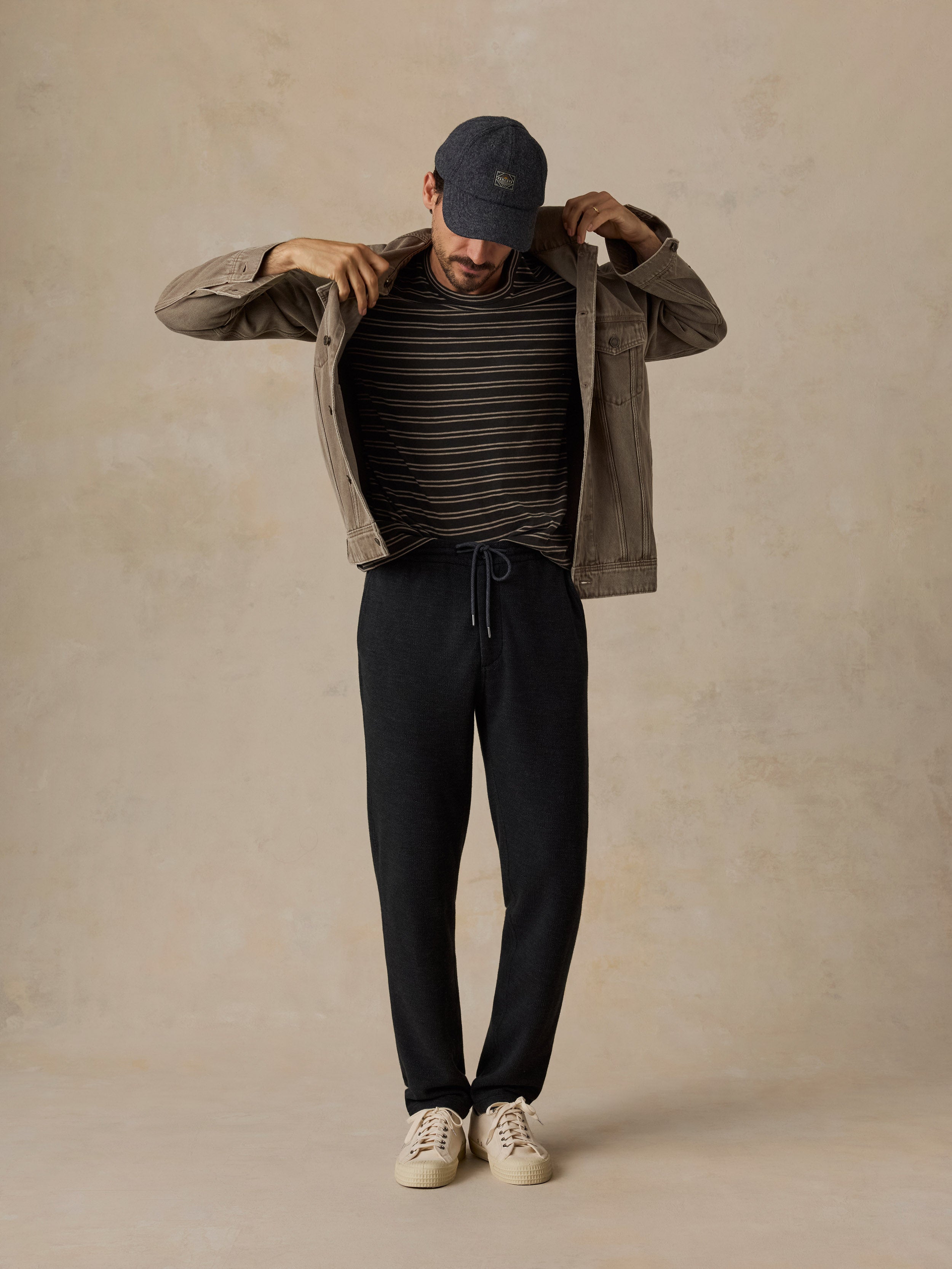 A man modeling a casual outfit consisting of a dark olive green utility jacket, a black and beige striped t-shirt, black sweatpants, a navy blue baseball cap, and off-white sneakers, set against a textured beige studio backdrop.