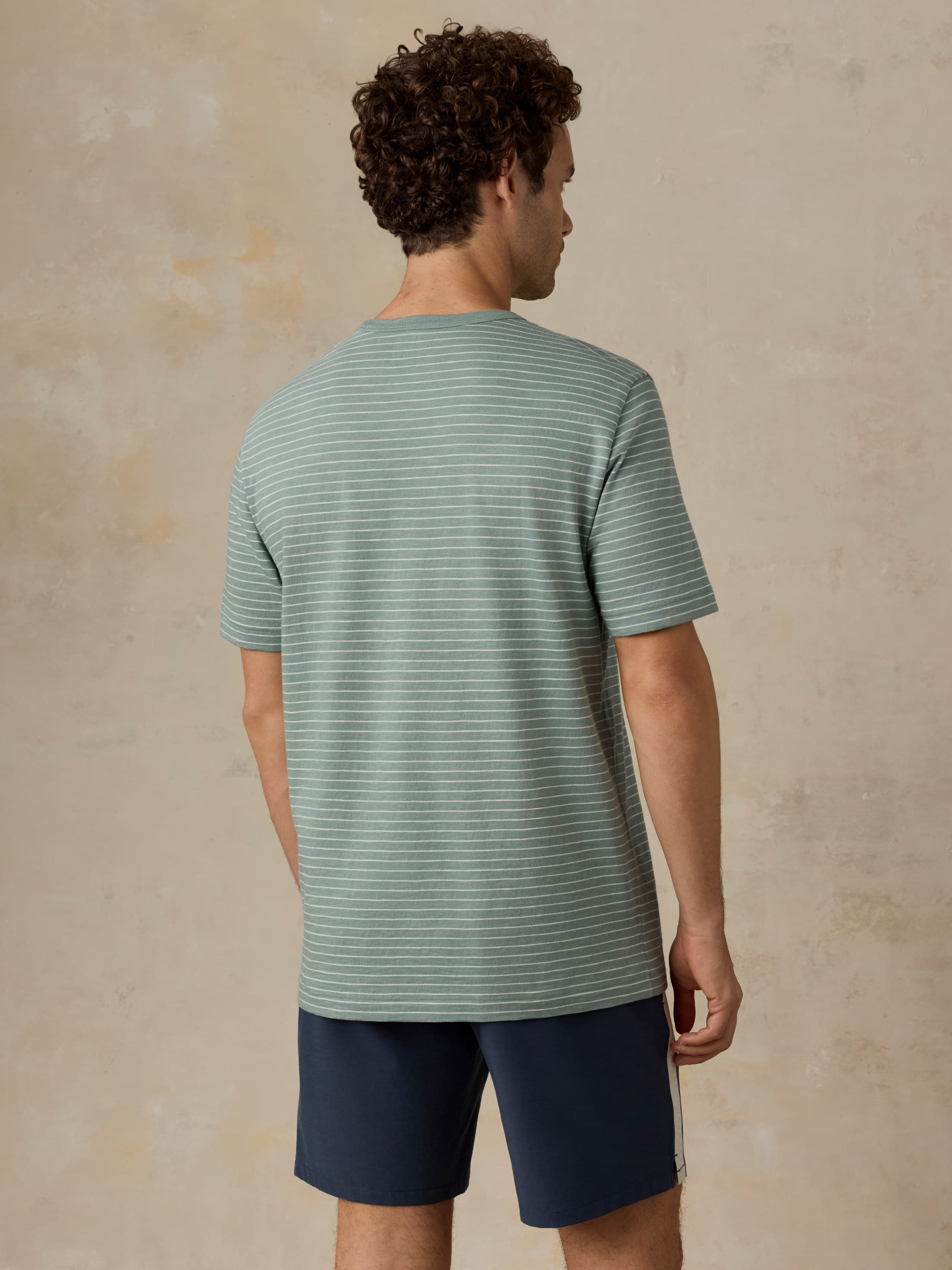 Man wearing a green and white striped tee with navy swim shorts against a beige background